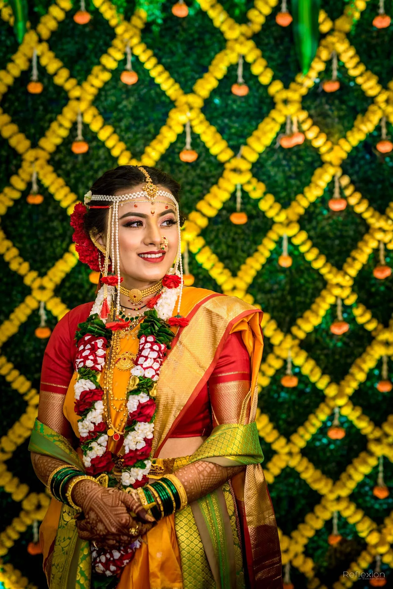A bride in traditional Indian wedding attire standing in front of a vibrant yellow and green floral backdrop with small orange lamps hanging.