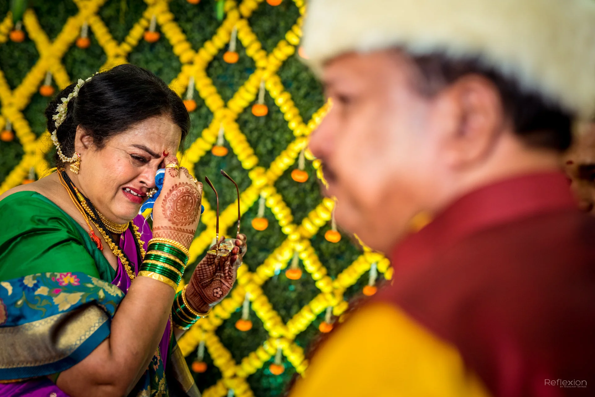 An older woman dressed in traditional Indian attire is crying and holding her face with one hand, while a man, also in traditional clothing, looks on with a serious expression. The background is decorated with yellow flowers and small orange decorati