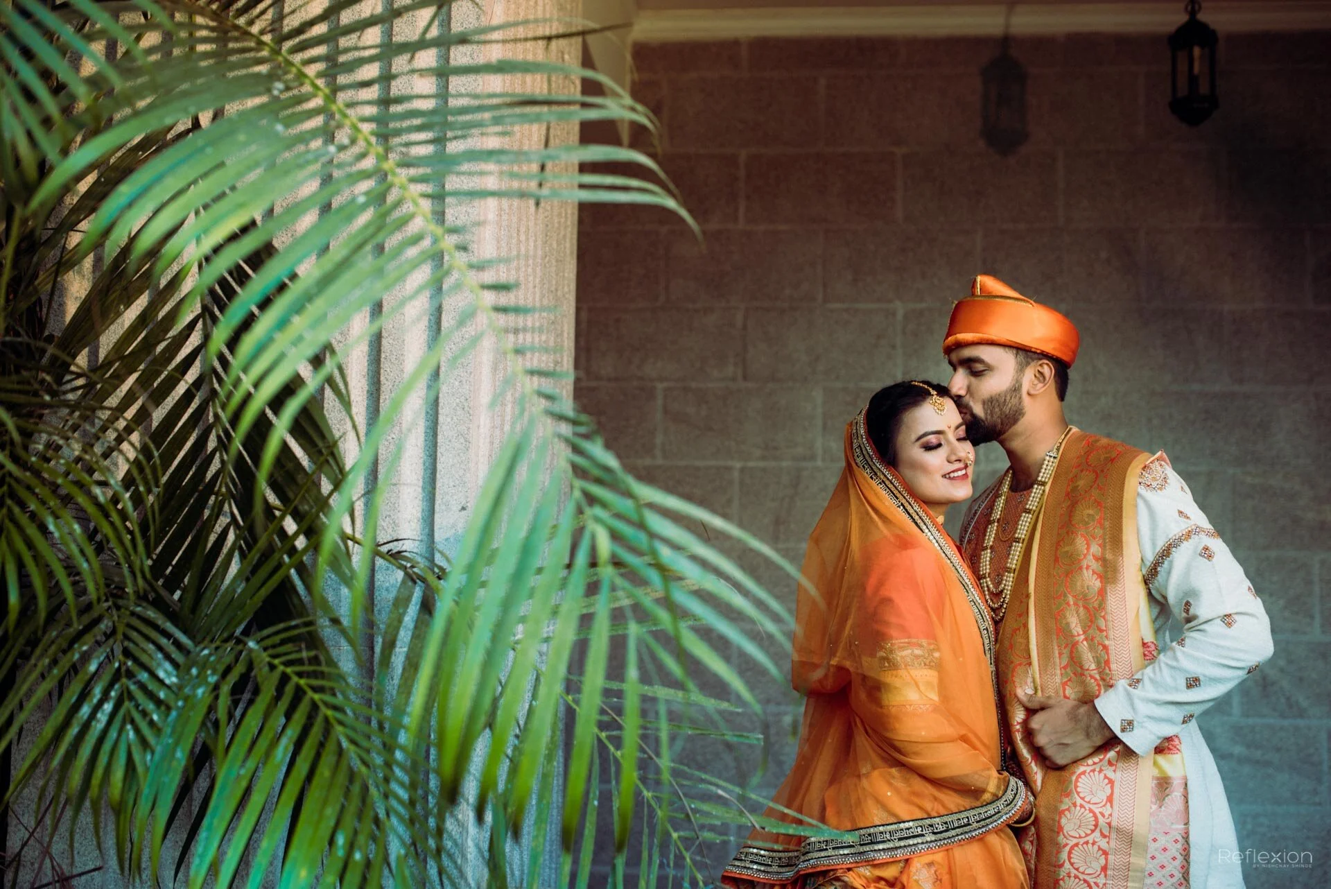 A newlywed Indian couple dressed in traditional attire, embracing in a warm moment, with lush green plants in the foreground and a brick wall background.