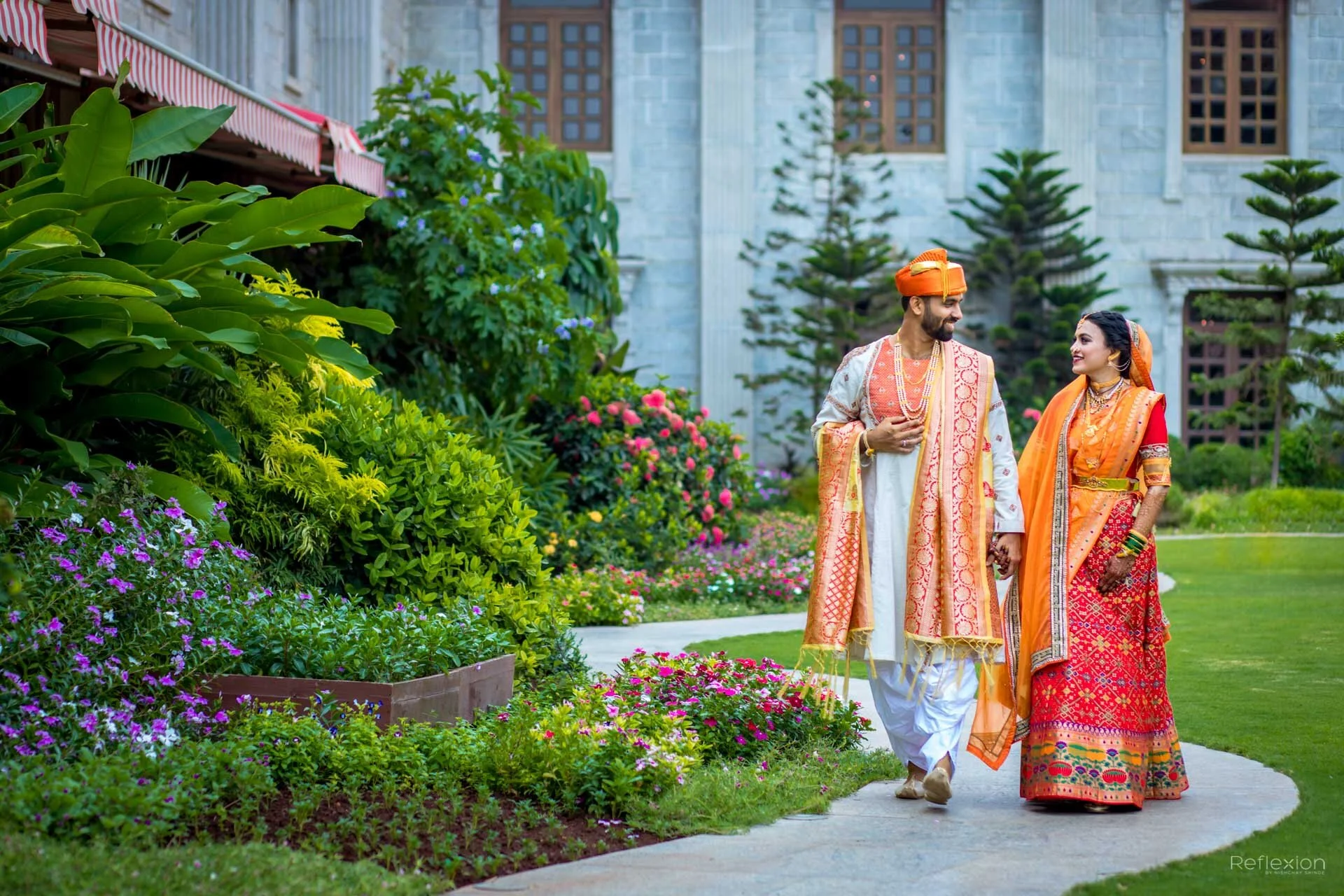 A couple dressed in traditional Indian wedding attire walking hand in hand on a garden path surrounded by colorful flowers and lush greenery, with a stone building in the background.