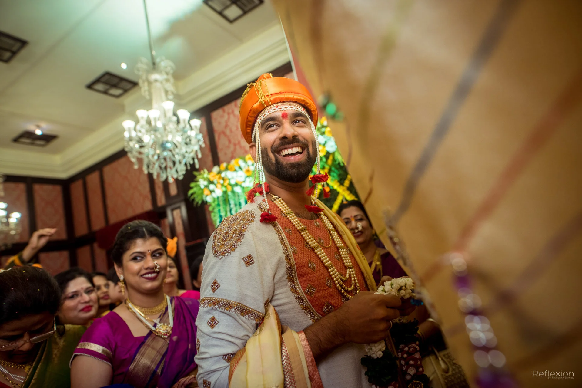 Happy groom in traditional Indian wedding attire holding a floral garland surrounded by smiling women at the celebration.