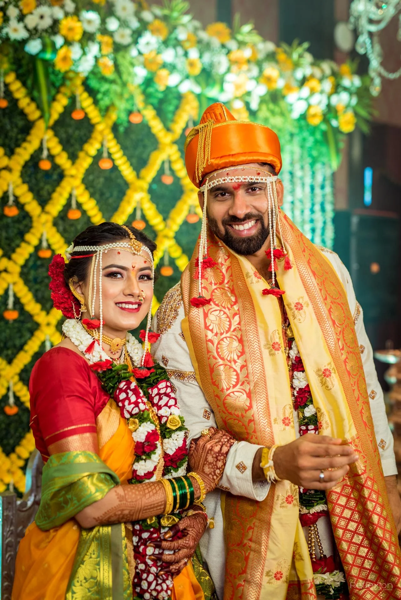A newlywed Indian couple dressed in traditional wedding attire, standing close together and smiling at the camera. The bride wears a red and gold saree with elaborate jewelry and a flower garland, while the groom wears a cream-colored sherwani with a