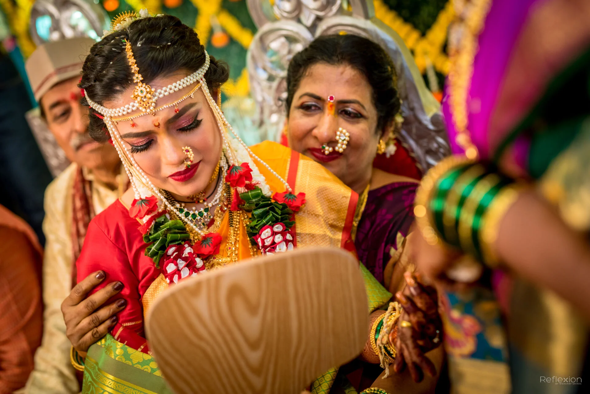 A bride in traditional Indian wedding attire is being presented with a wooden item by an older woman. The bride is wearing a bright saree, heavy jewelry, and floral necklaces, and appears emotional. The scene is decorated with colorful flowers and th