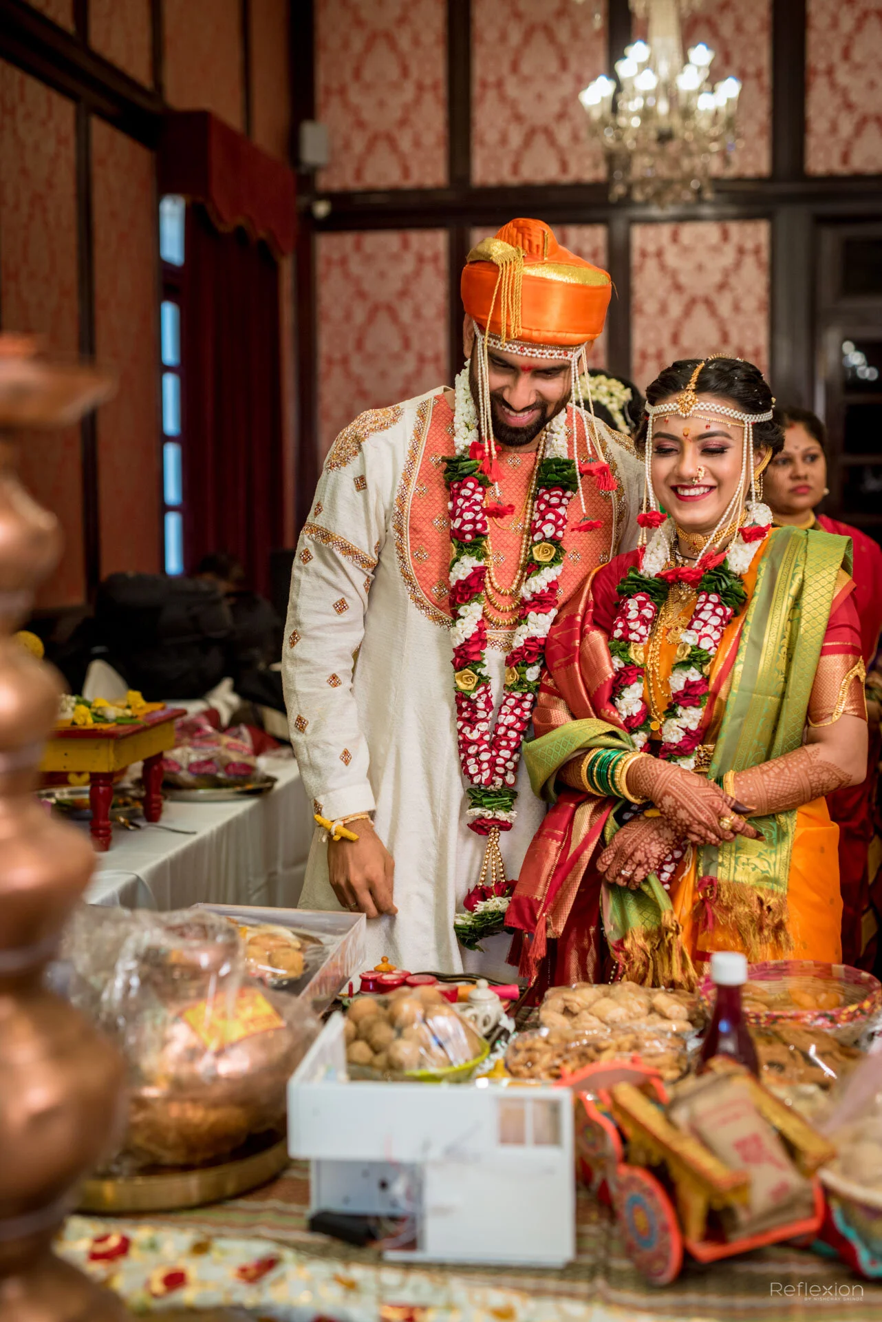 Indian wedding ceremony with bride and groom wearing traditional attire, garlands, and jewelry, standing beside each other and smiling, surrounded by offerings and decorations.