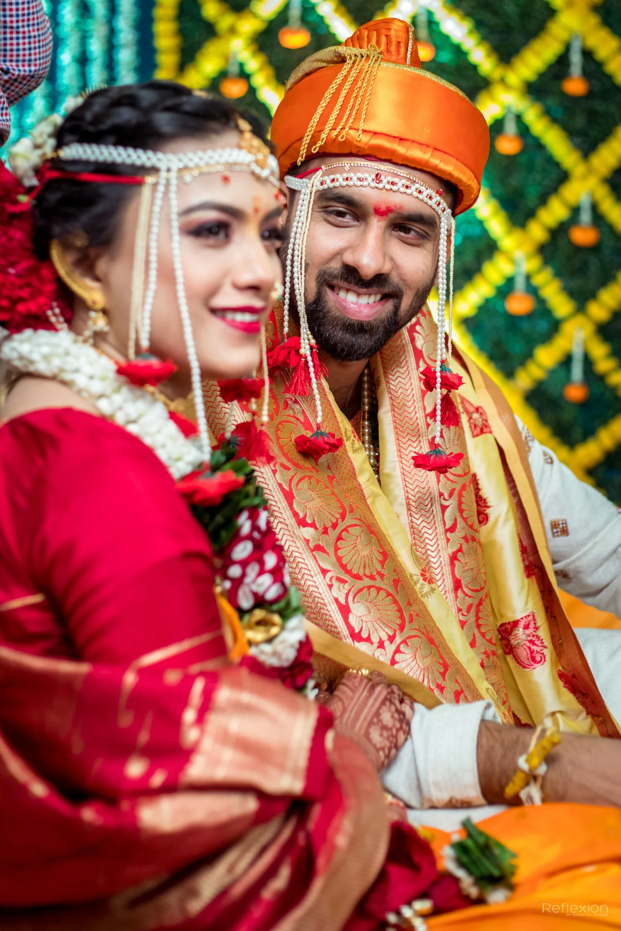 A couple in traditional Indian wedding attire, sitting together during a celebration with colorful decorations in the background.