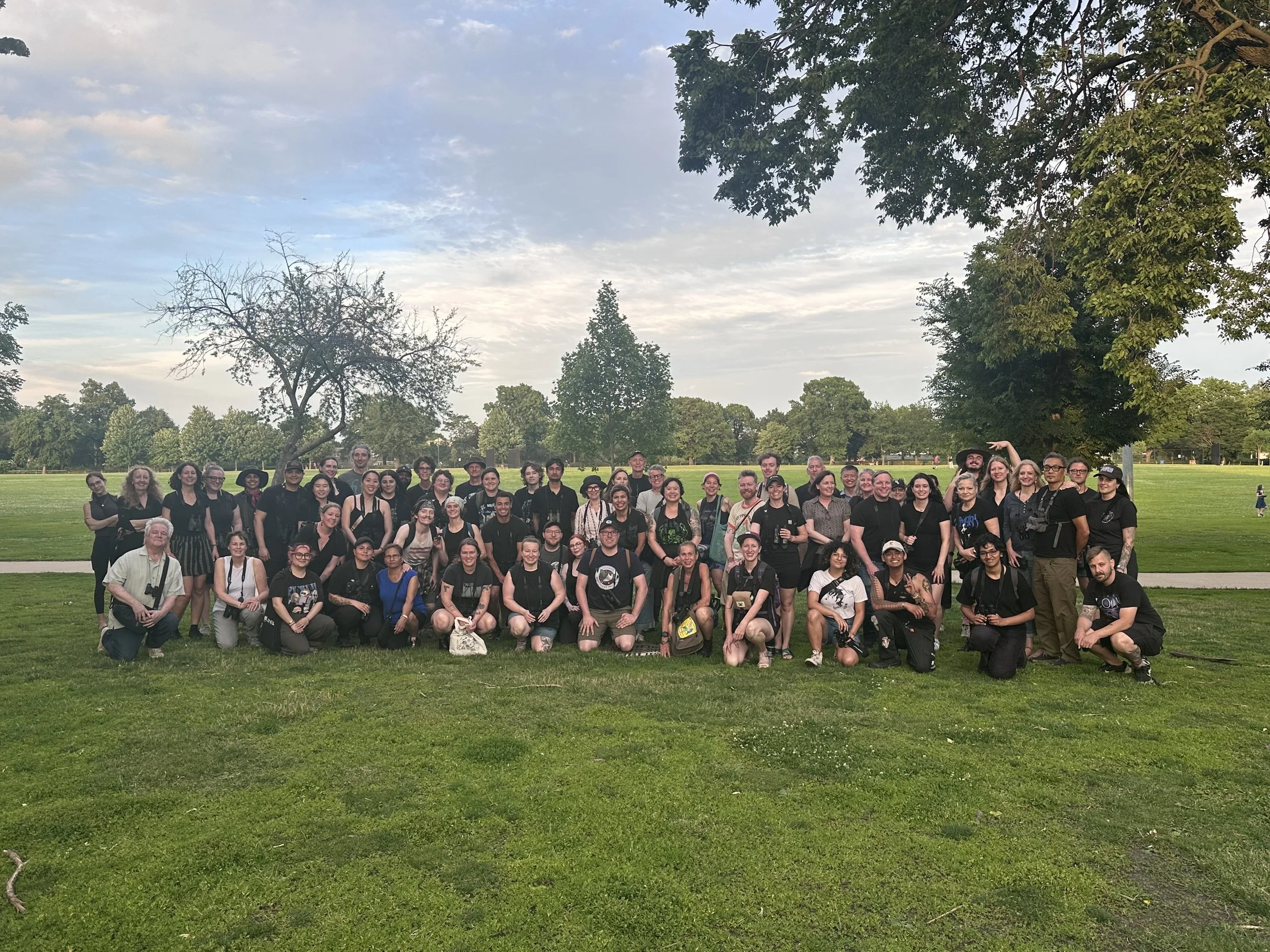 About 70 people, most in dark lightweight clothes, face the camera in two rows, the front row on their knees. They are on a grassy lawn with a few trees and a sidewalk behind them.