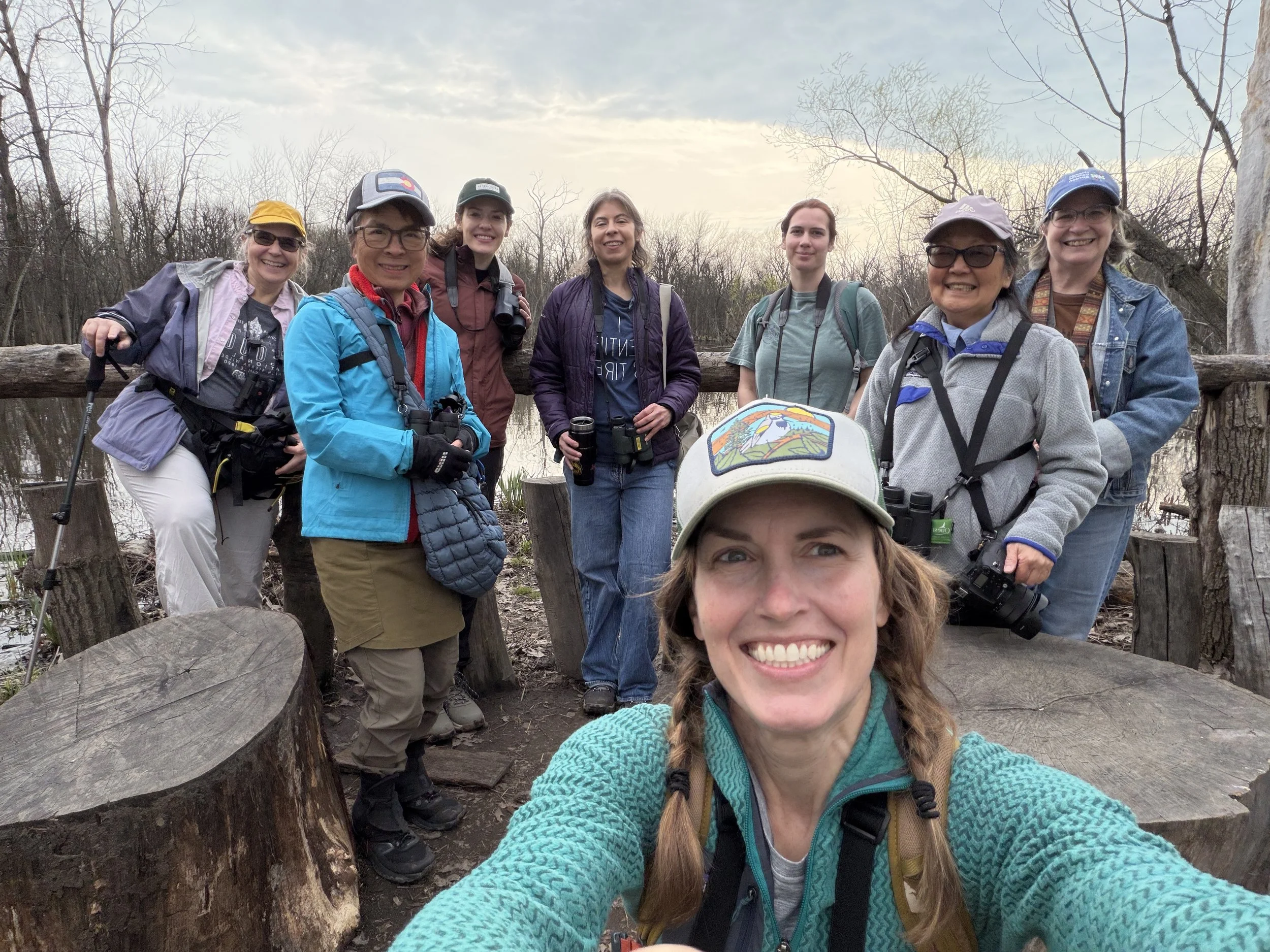 Eight people in jackets, with binoculars, smiling at camera in front of a wooden railing with the slough behind.