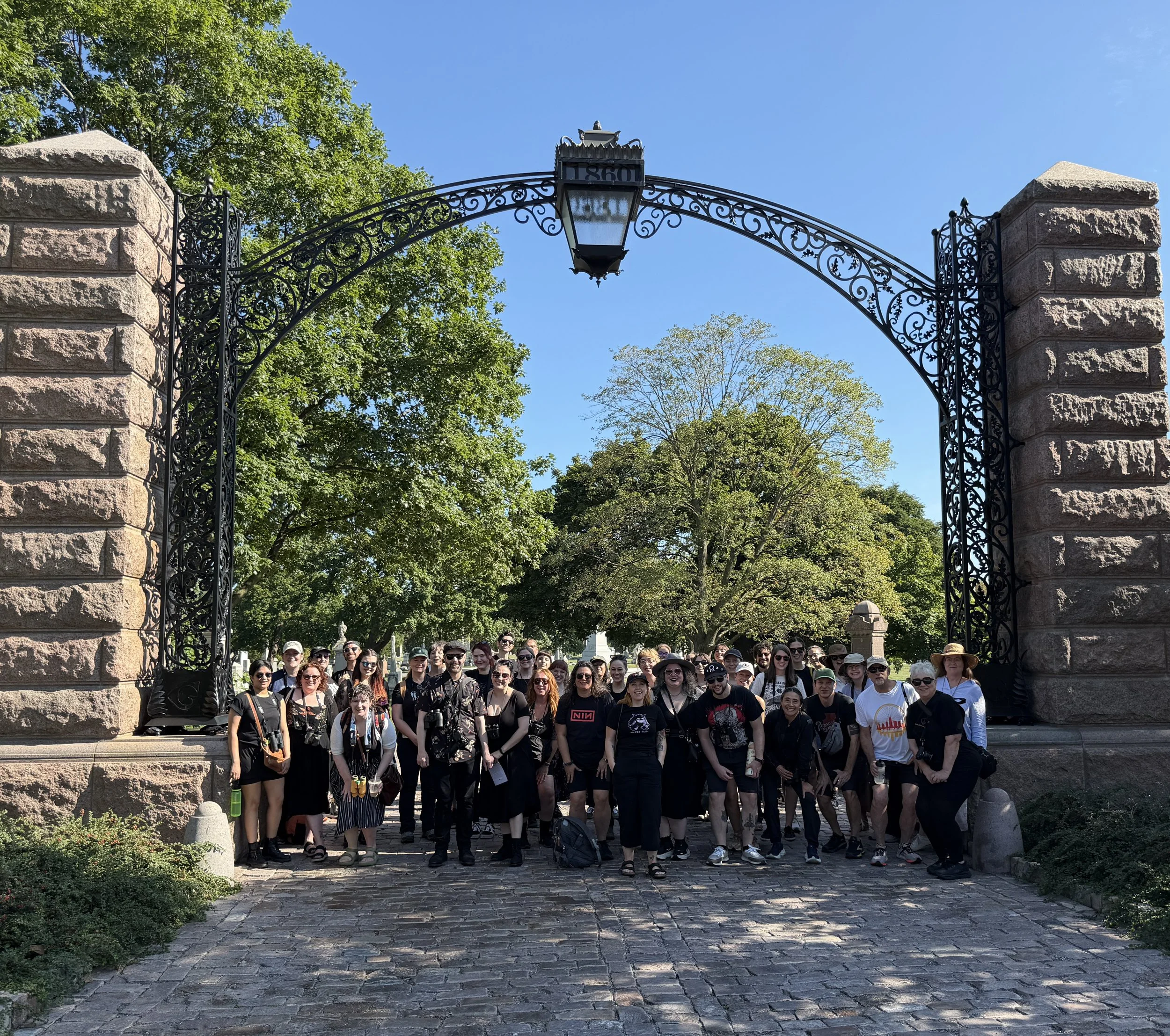 About 40 people facing camera, looking small under high wrought iron arch flanked by stone pillars, smiling, with green trees and bkue sky behind.