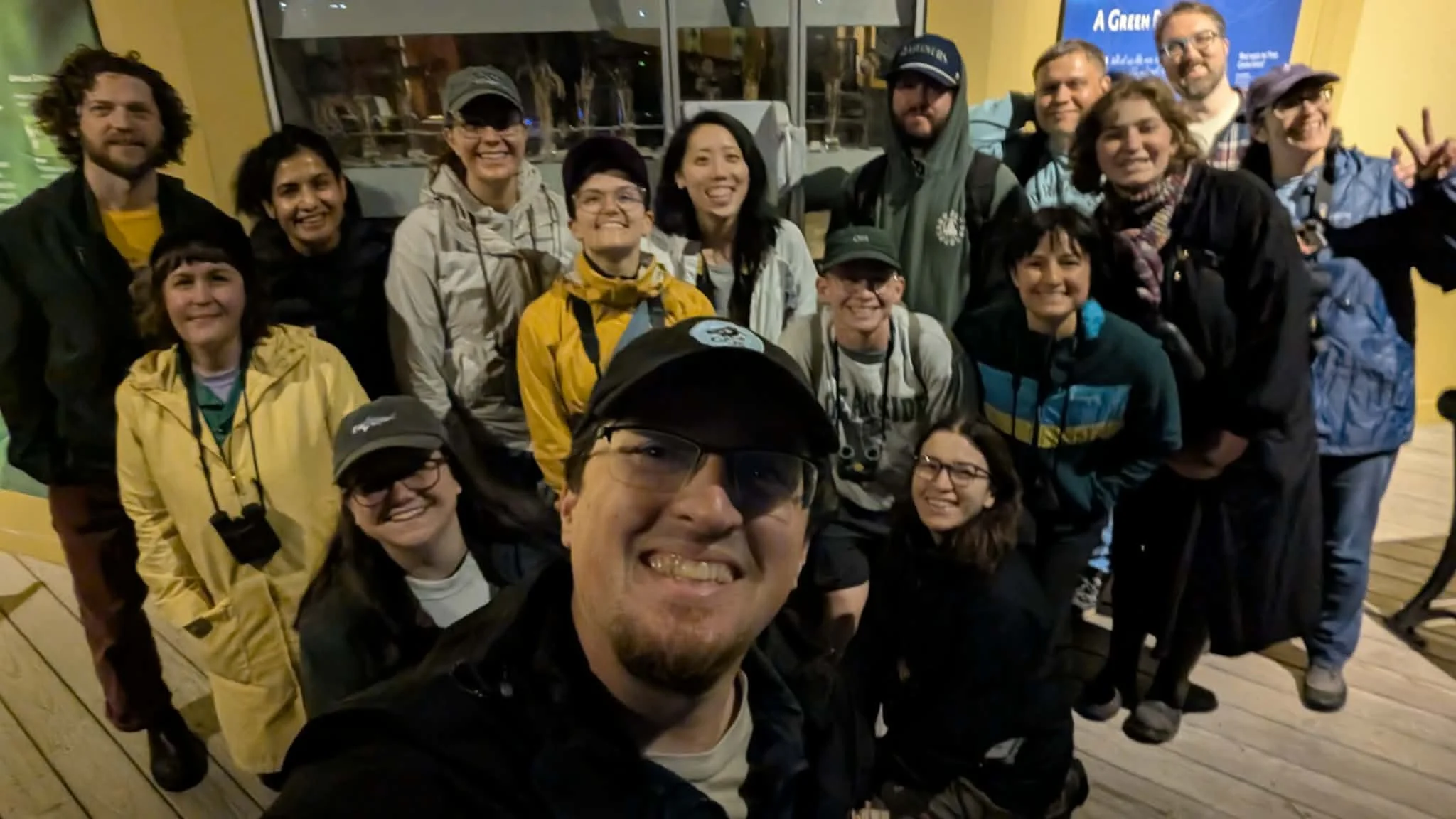 Fifteen people in jackets, with binoculars, smiling at camera in front of glass doors to nature center.