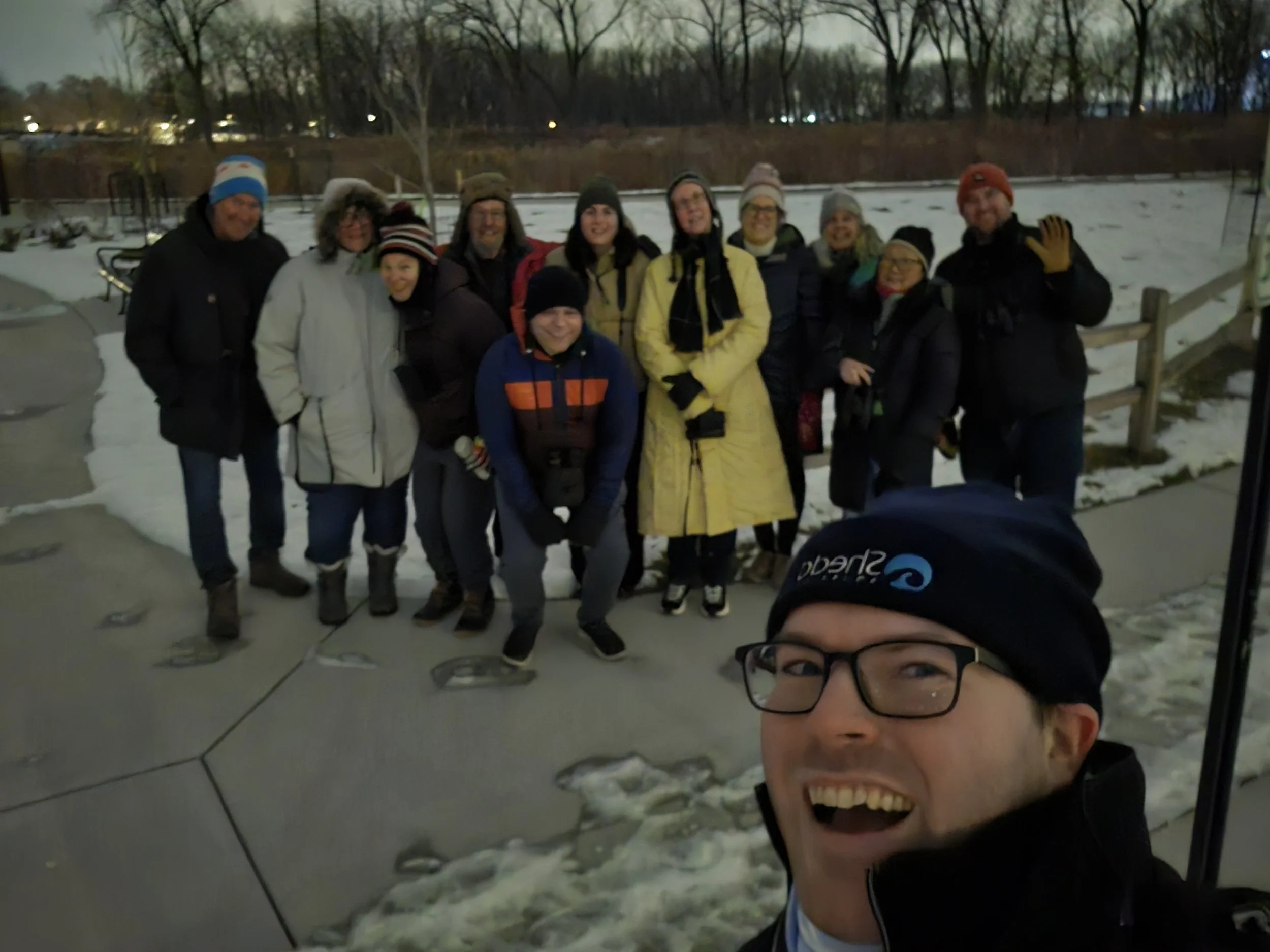 Ten people bundled in winter clothing in front of snowy field and distant trees at sunset