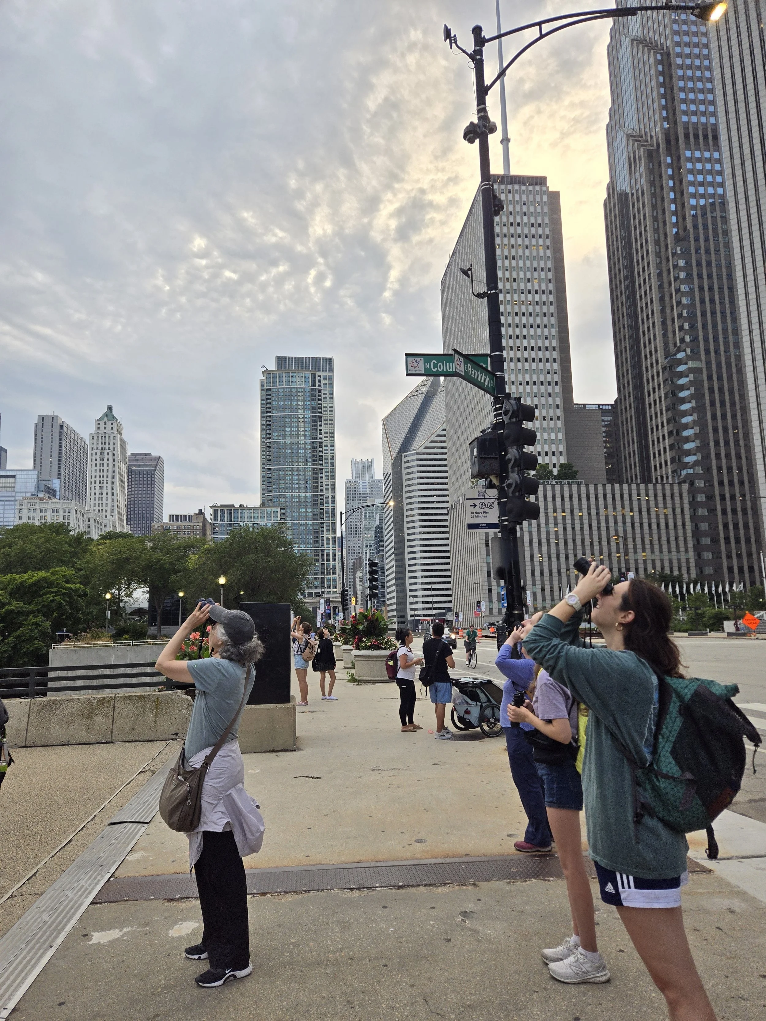 People with binoculars peering into the sky against a backdrop of skyscrapers and commuters.