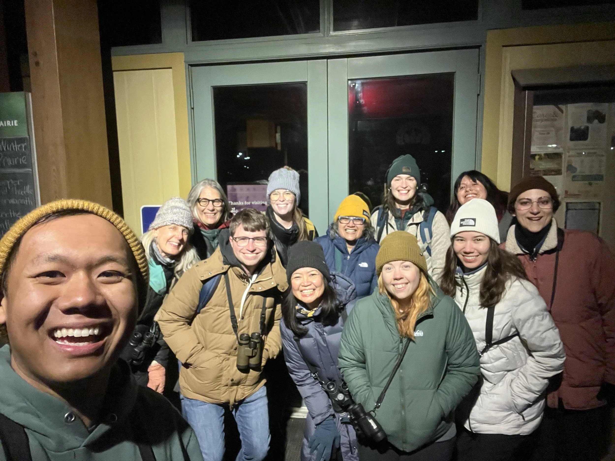 Twelve people in parkas and hats, smiling at the camera in front of the doors to a small building.