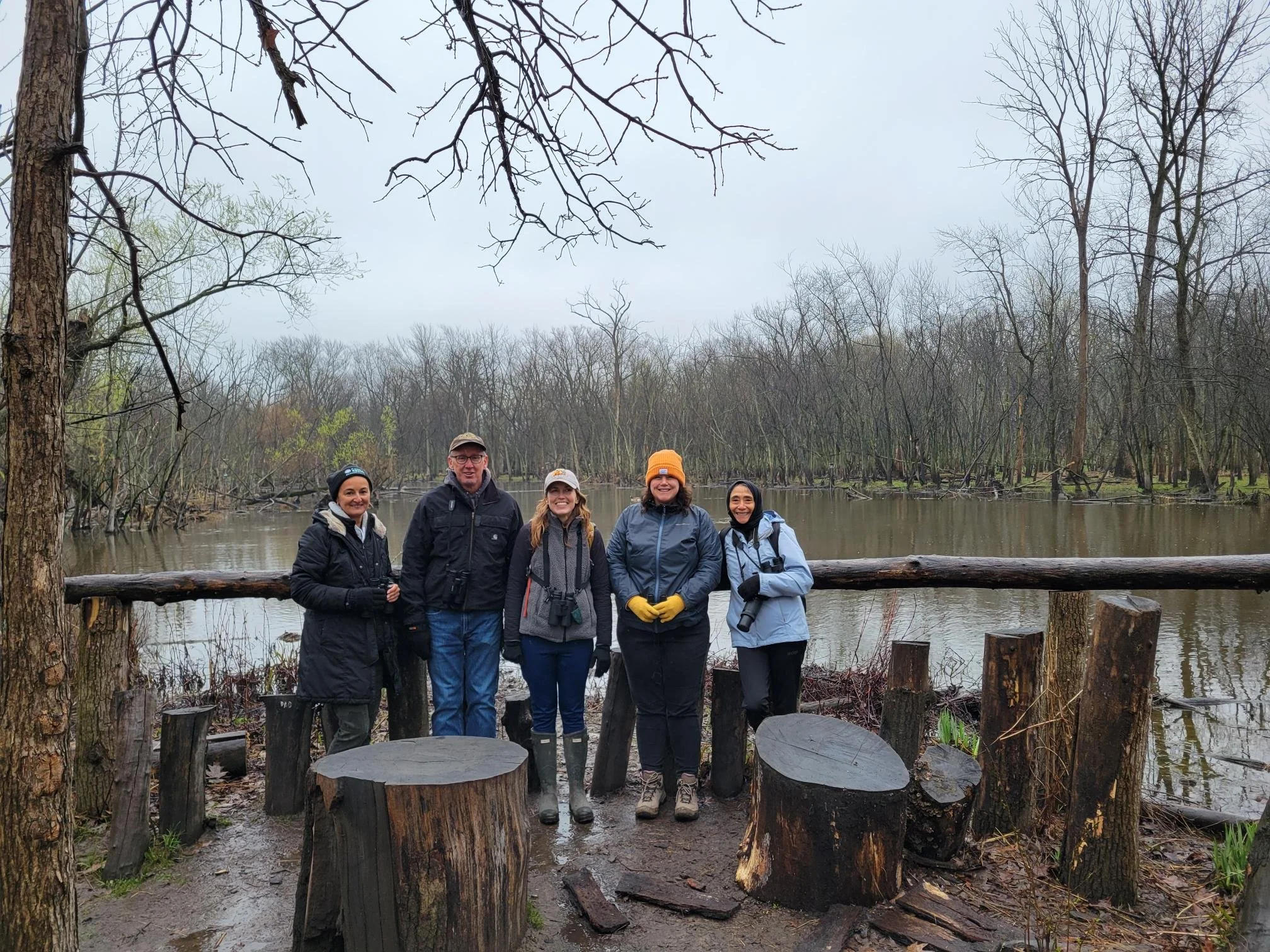Five people in rain gear, with binoculars, face the camera, smiling. There is a wooden railing behind them and behind that a lagoon. Leafless trees are in the background.