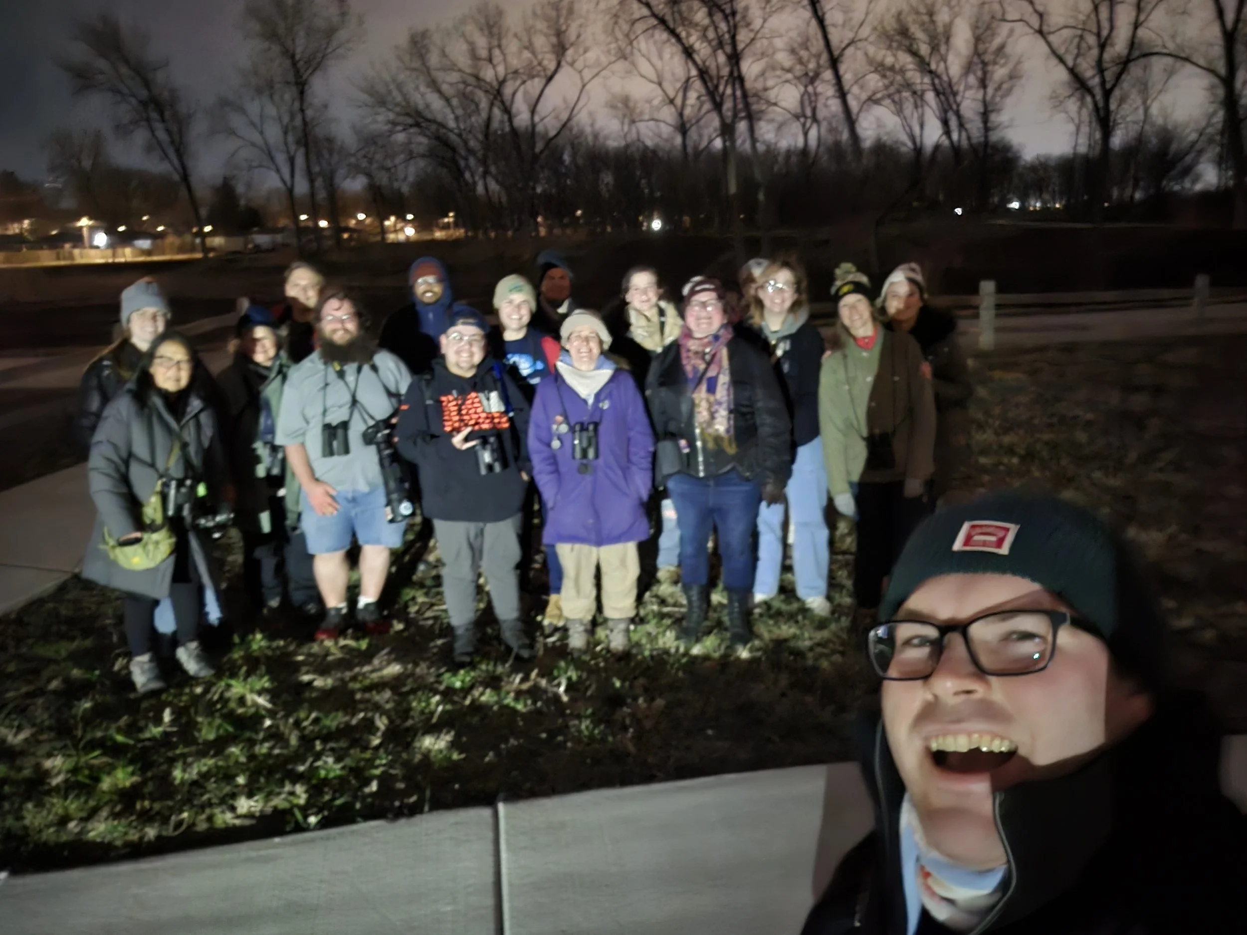 19 people dressed warmly with binoculars smiling at the camera, standing on grass near sidewalk, dark sky and leafless trees behind them.