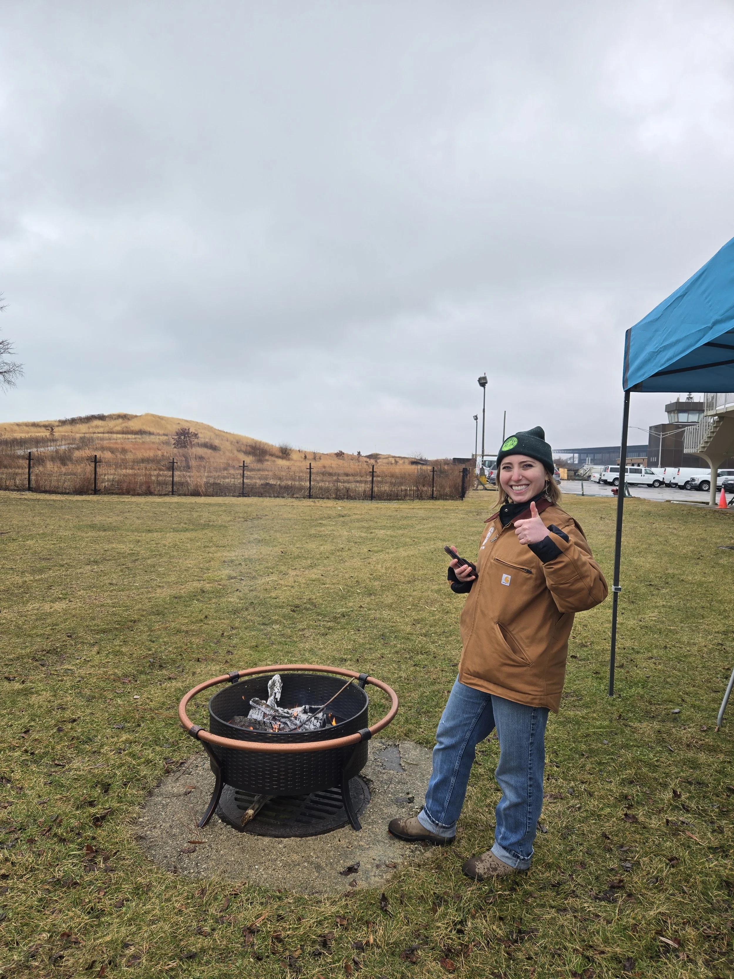 Smiling woman in heavy coat and knit cap gives a thumbs up as she stokes a fire pit, with the Northerly Island hill behind her.