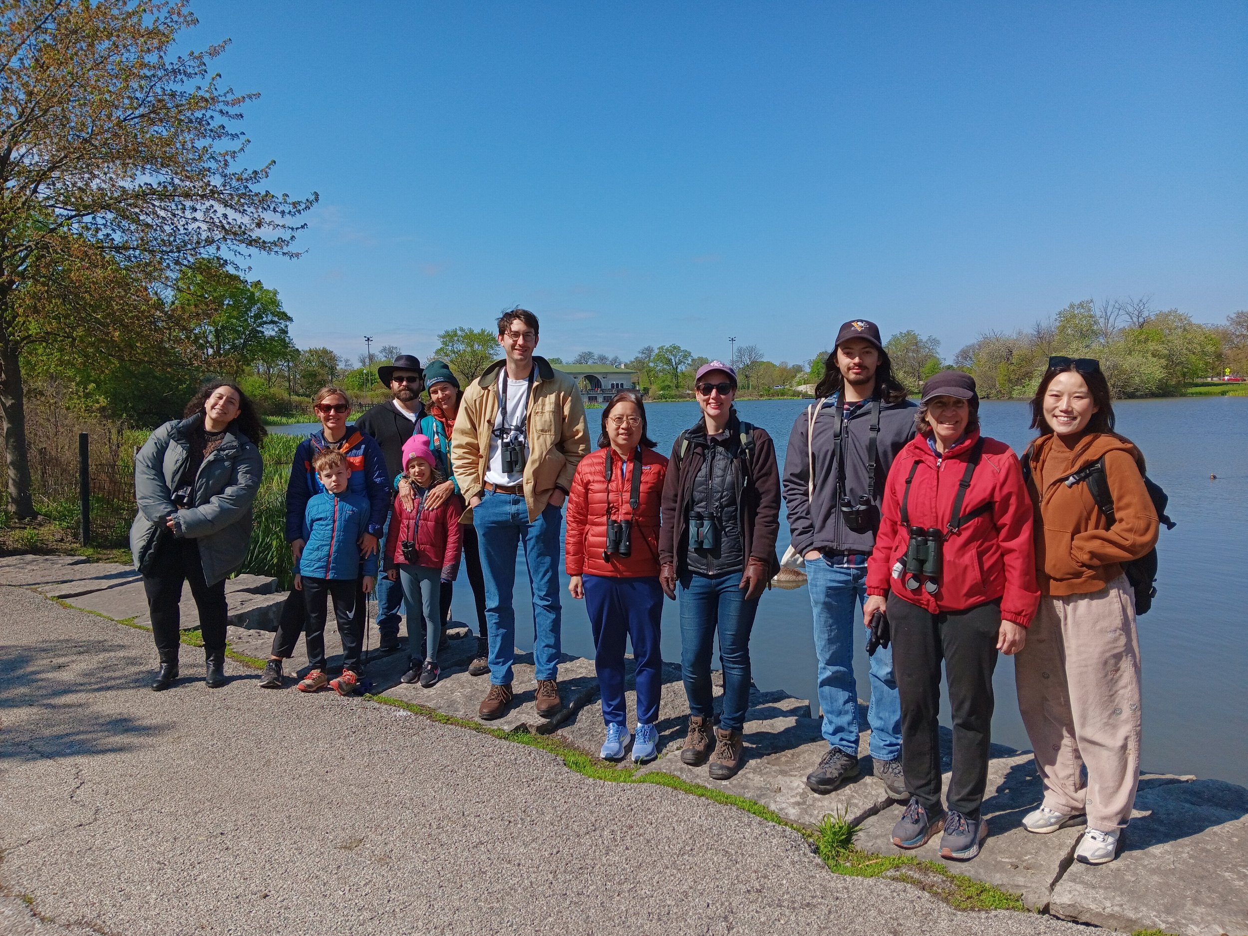 Twelve people, including two children, in jackets and with binoculars on sidewalk with lagoon behind them