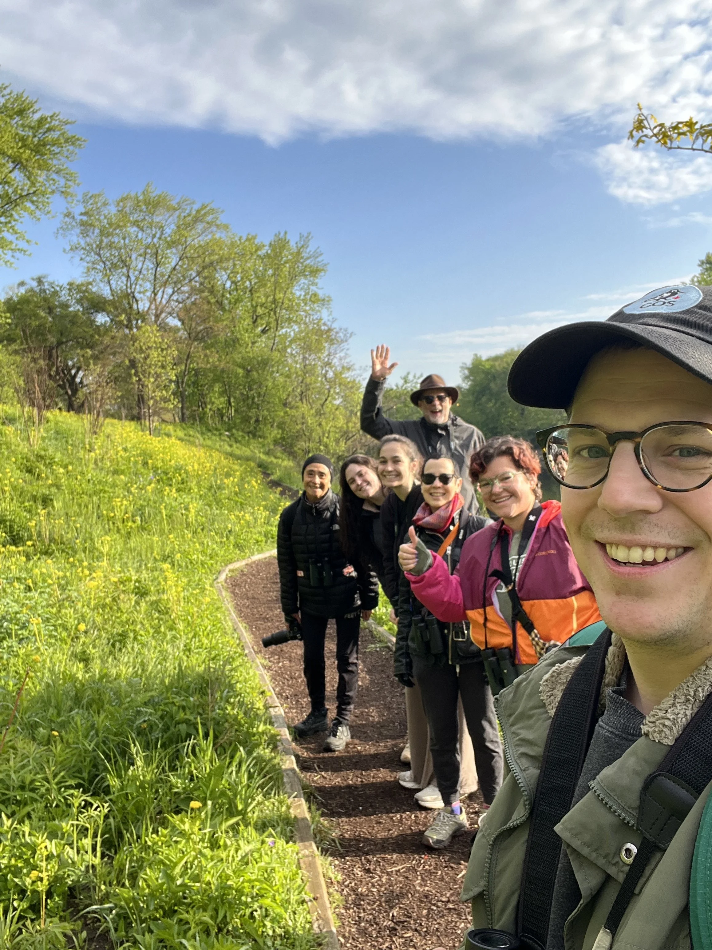 Ten people in a row on the right side of the photo, facing camera, waving or thumbs up, along a wood chip path with long green grasses on the left side and trees in the back.