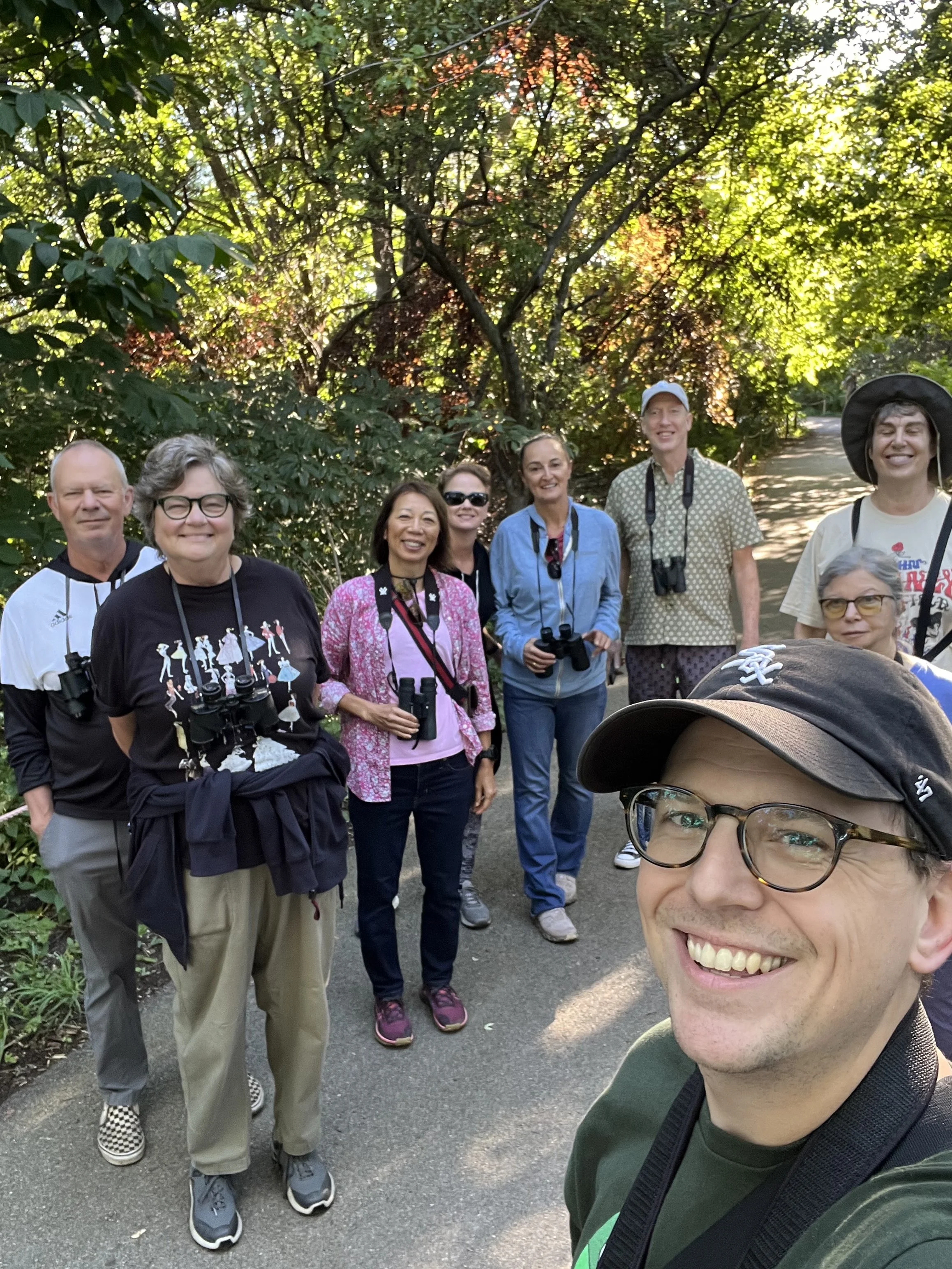 Eight people facing camera, smiling, binoculars around their necks, with trees with green and yellow leaves behind them