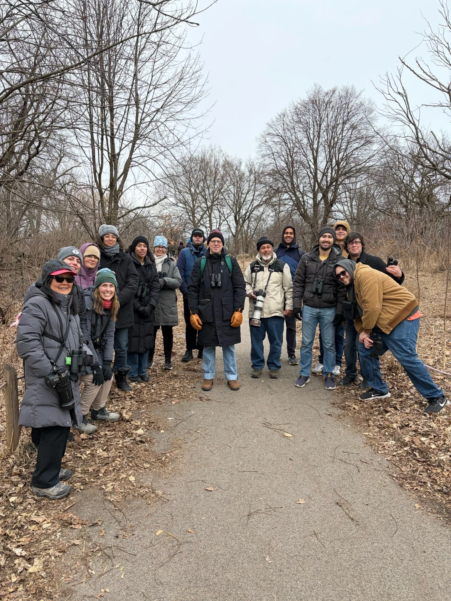 Fifteen people in winter clothes, with binoculars, smile at the camera along a paved path with leafless trees in the background.