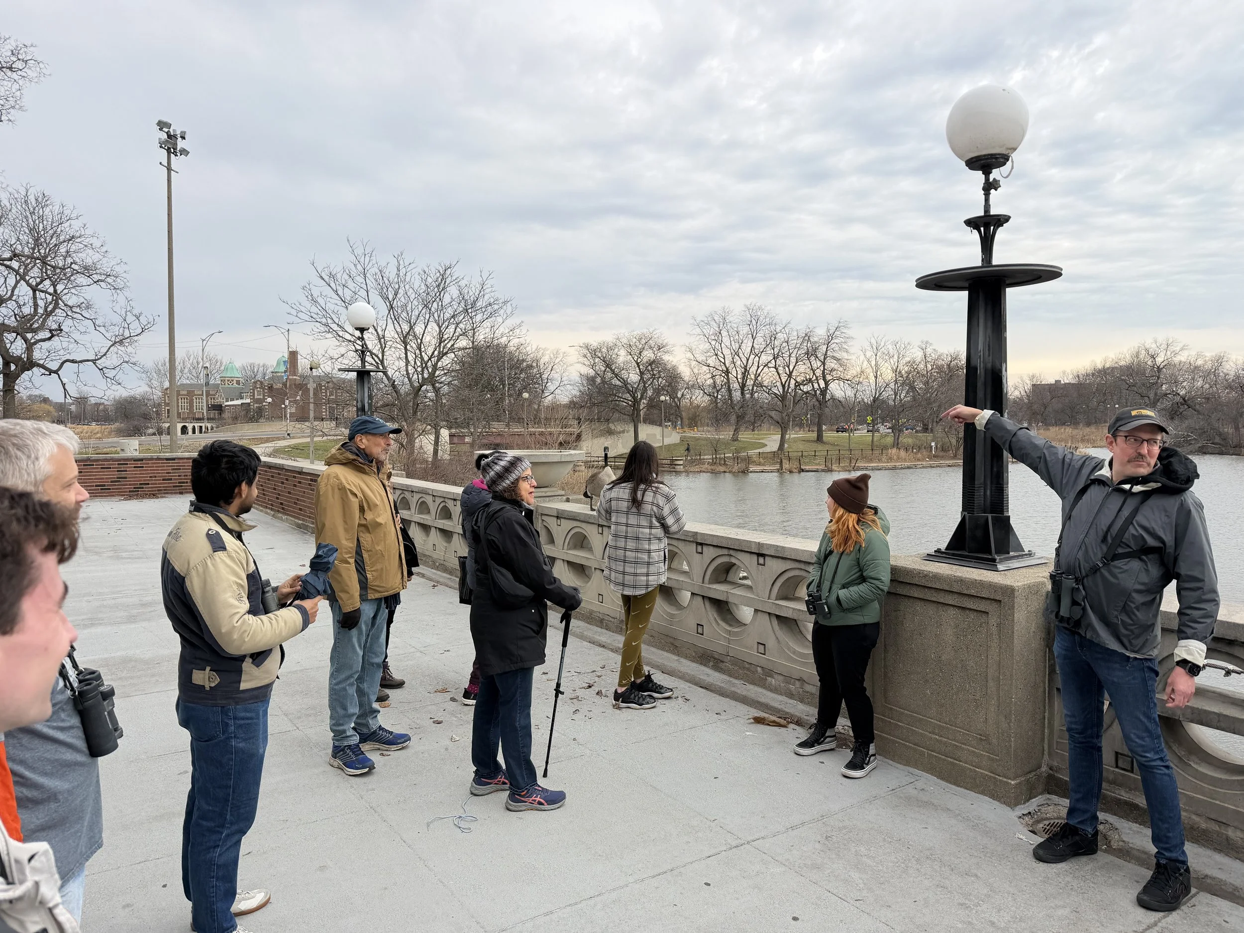 Man at right points to large gray and white goose on cement railing at edge of lagoon while seven people look on.