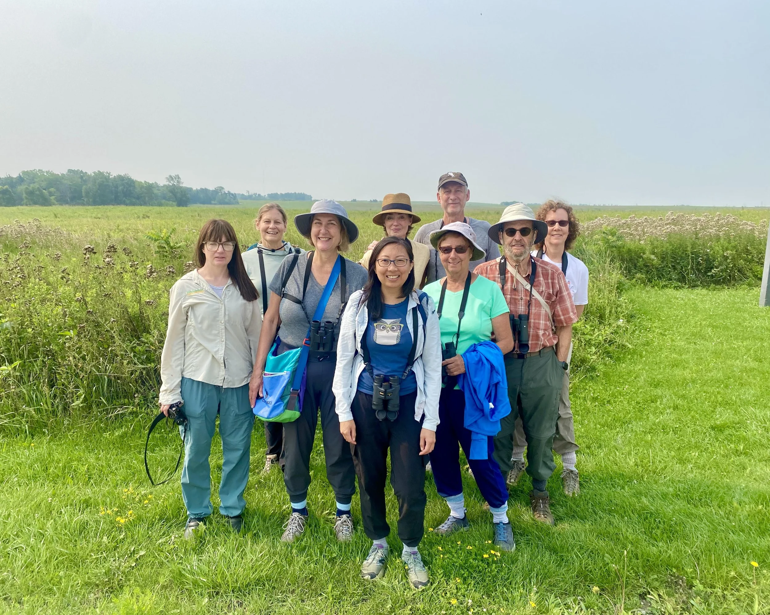 A group of 9 people with binoculars, some wearing hats and sunglasses, pose in front of a field of wild grasses. Behind them is a hazy blue-gray sky.