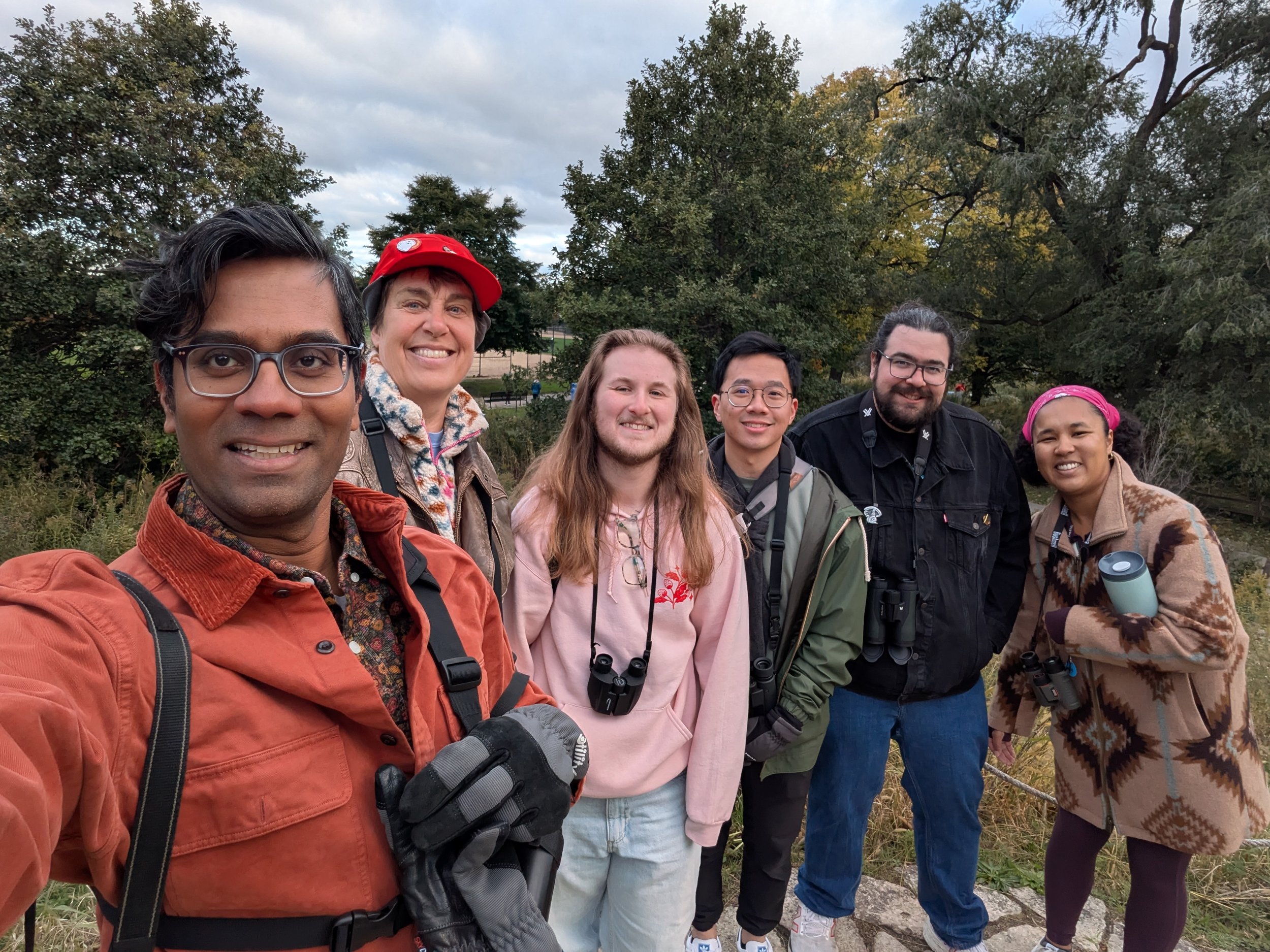 Six people in jackets and sweatshirts, with binoculars, smiling at the camera with trees and a partly cloudy sky behind.