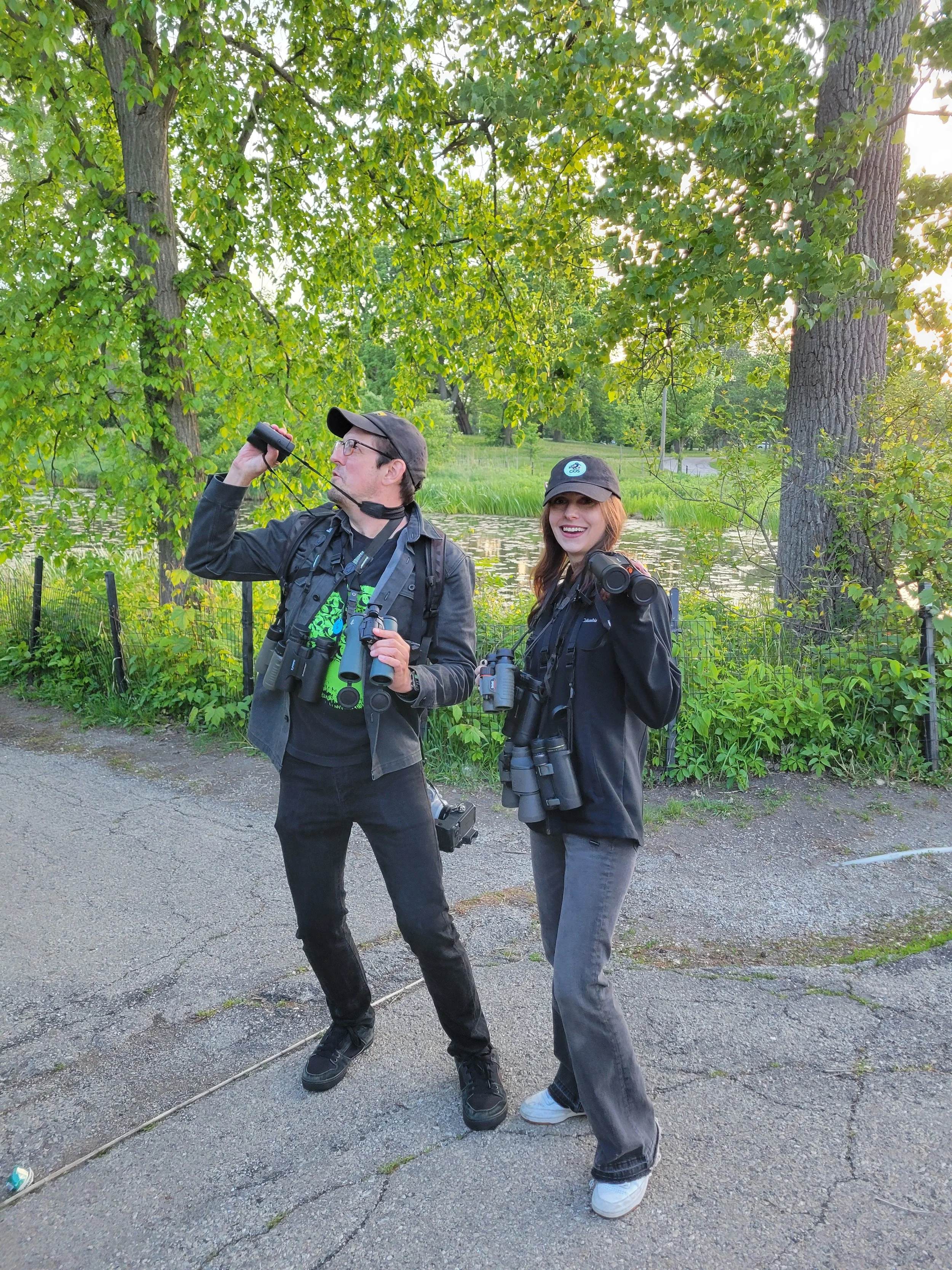 Two people with straps for multiple pairs of binoculars around their necks, one miming looking into the sky, the other laughing at the camera, with the lagoon and green shrubs behind them.