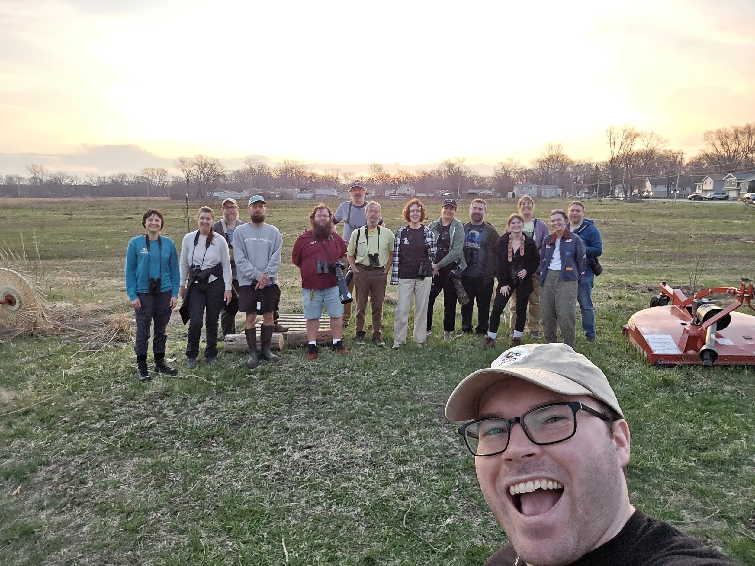 Fifteen people, one in foreground, smile at camera on a grassy field with setting sun behind them.