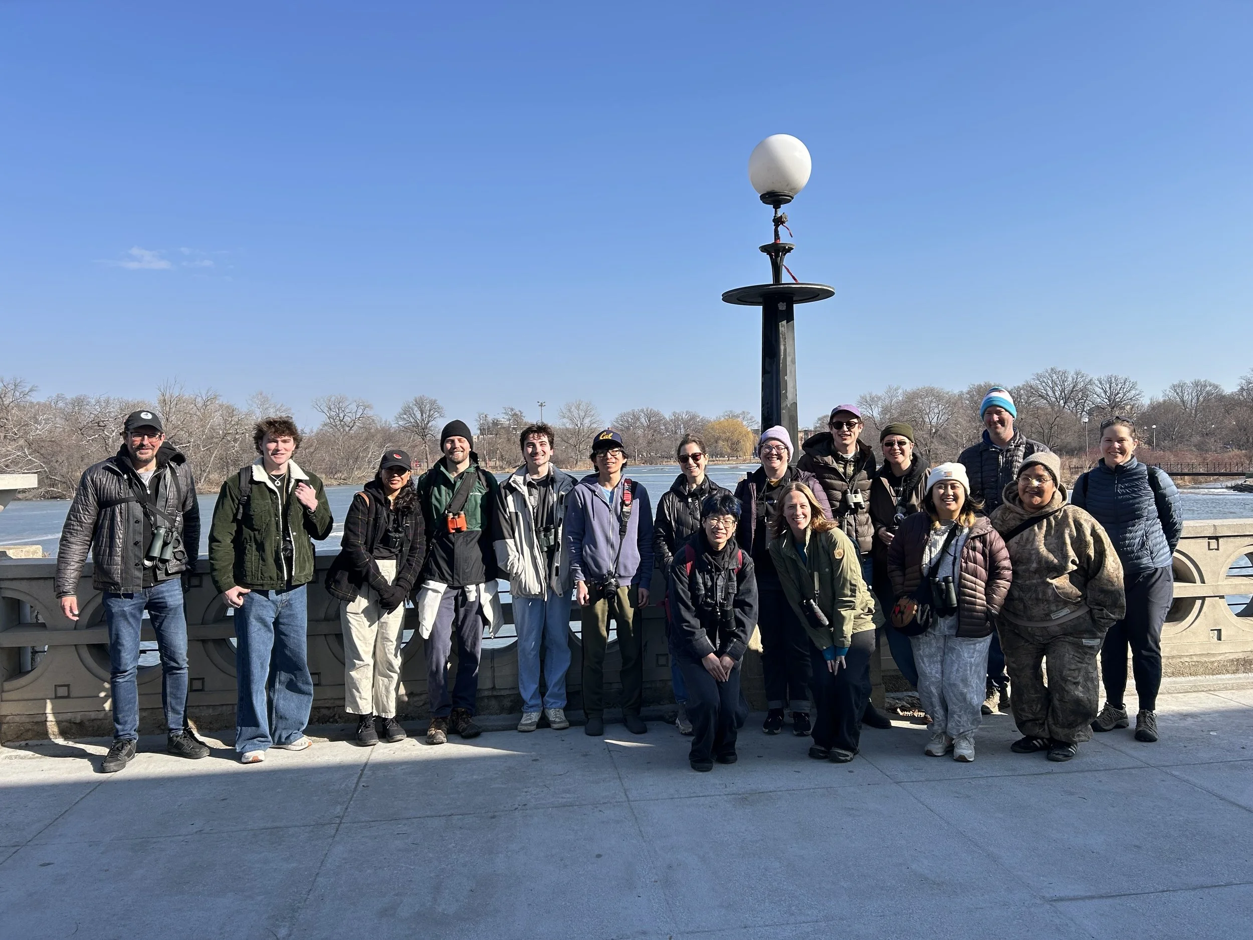 Seventeen people in winter gear, with binoculars, smile toward the camera with their backs against the stone wall in front of the lagoon.