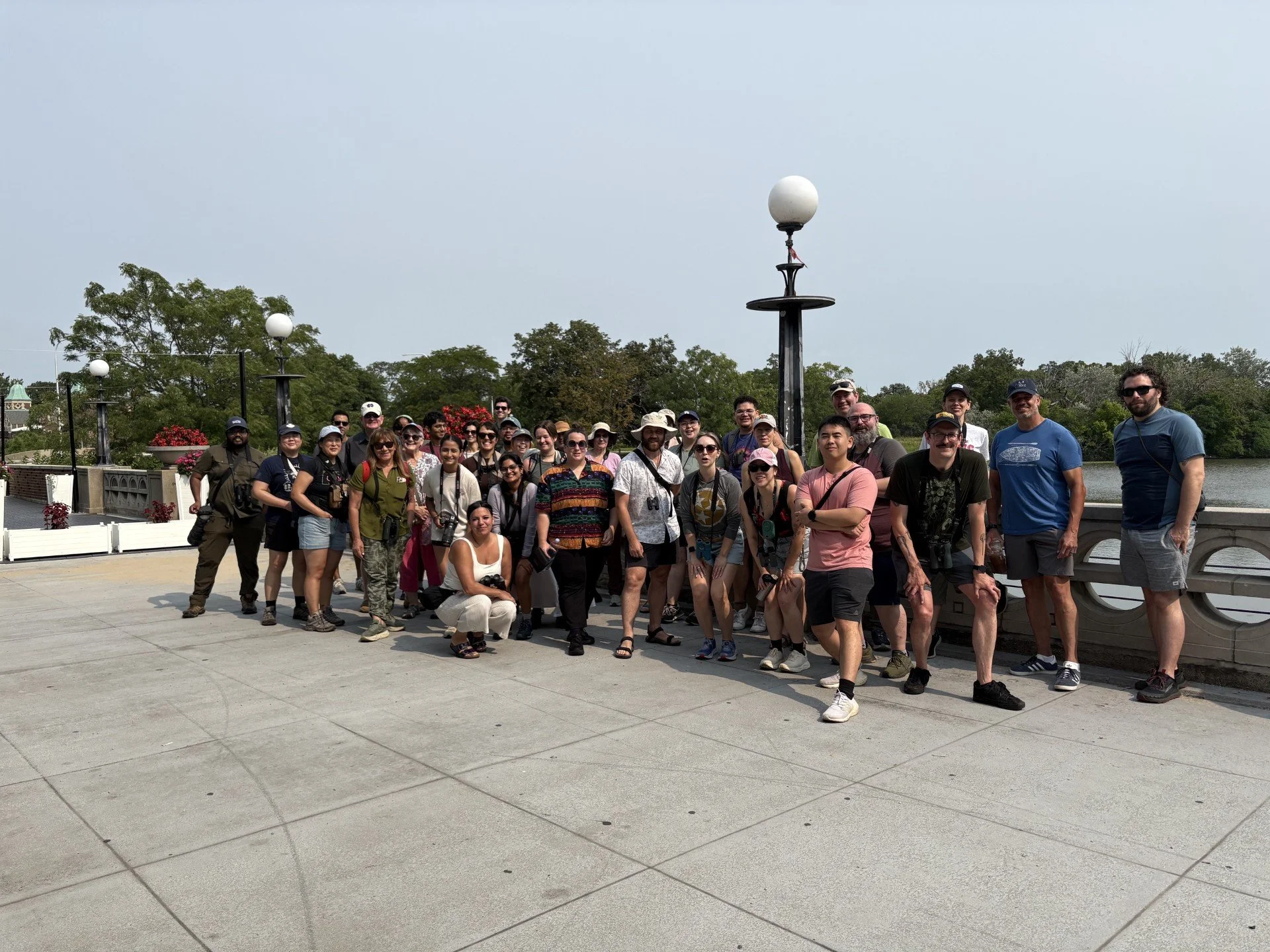 About 50 people, tightly grouped, wearing shorts and tshirts, smiling toward the camera with a concrete railing and the lagoon behind them.