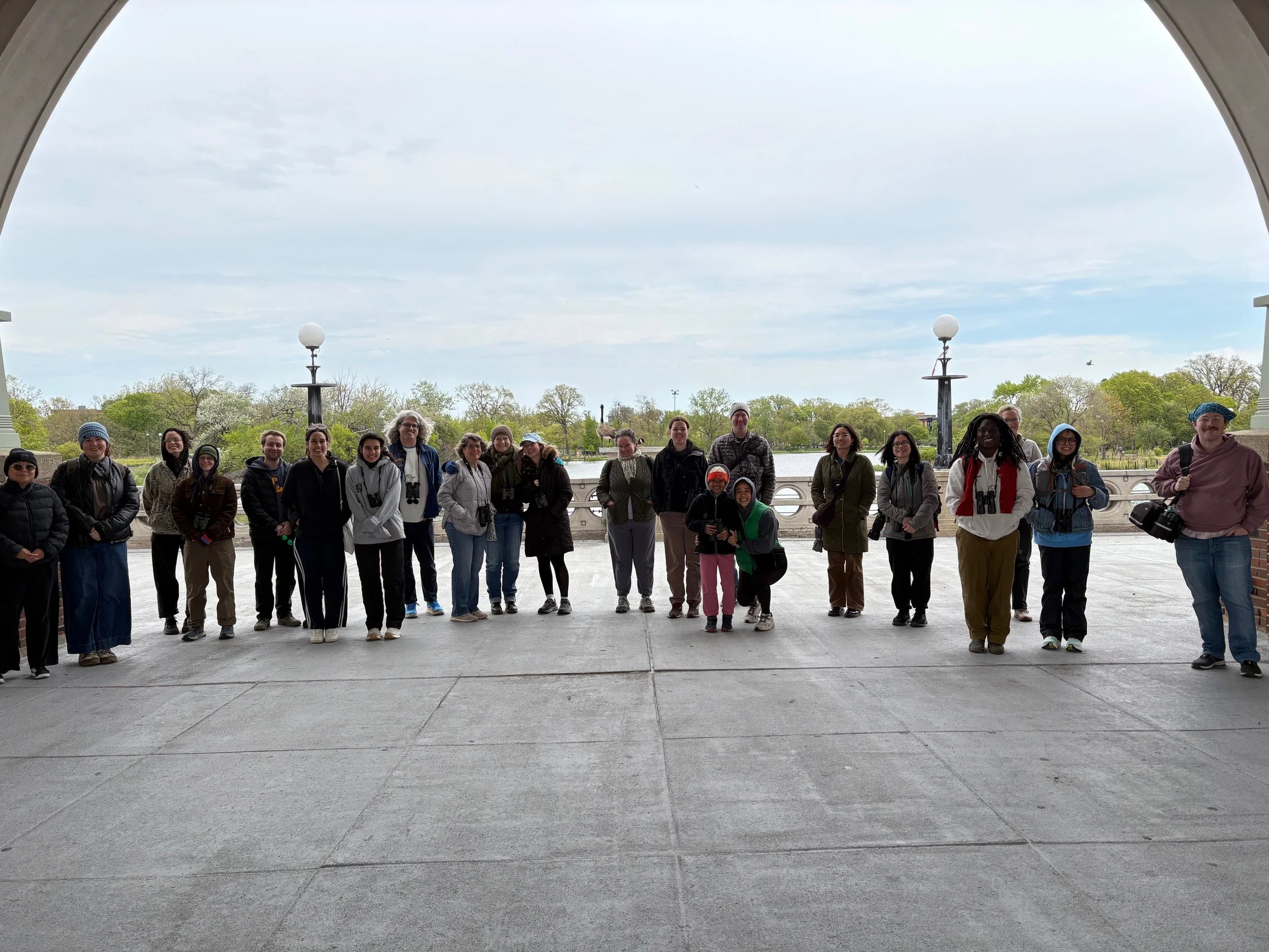 22 people in a line in warm coats, with binoculars, smiling toward camera on fieldhouse plaza, green trees behind them.