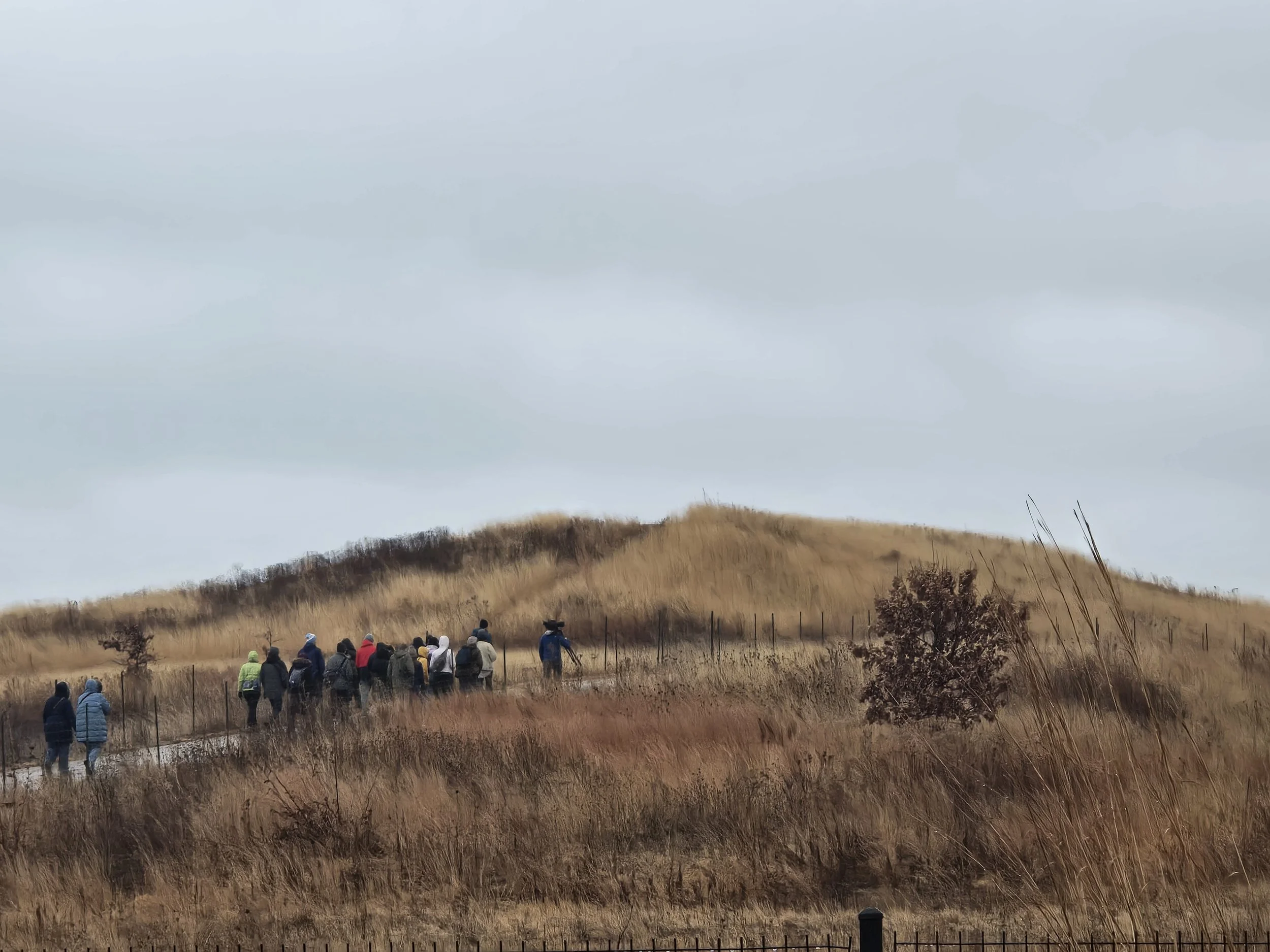 Sixteen people in winter gear, facing away from camera, hike up a low hill covered with brown winter grass.