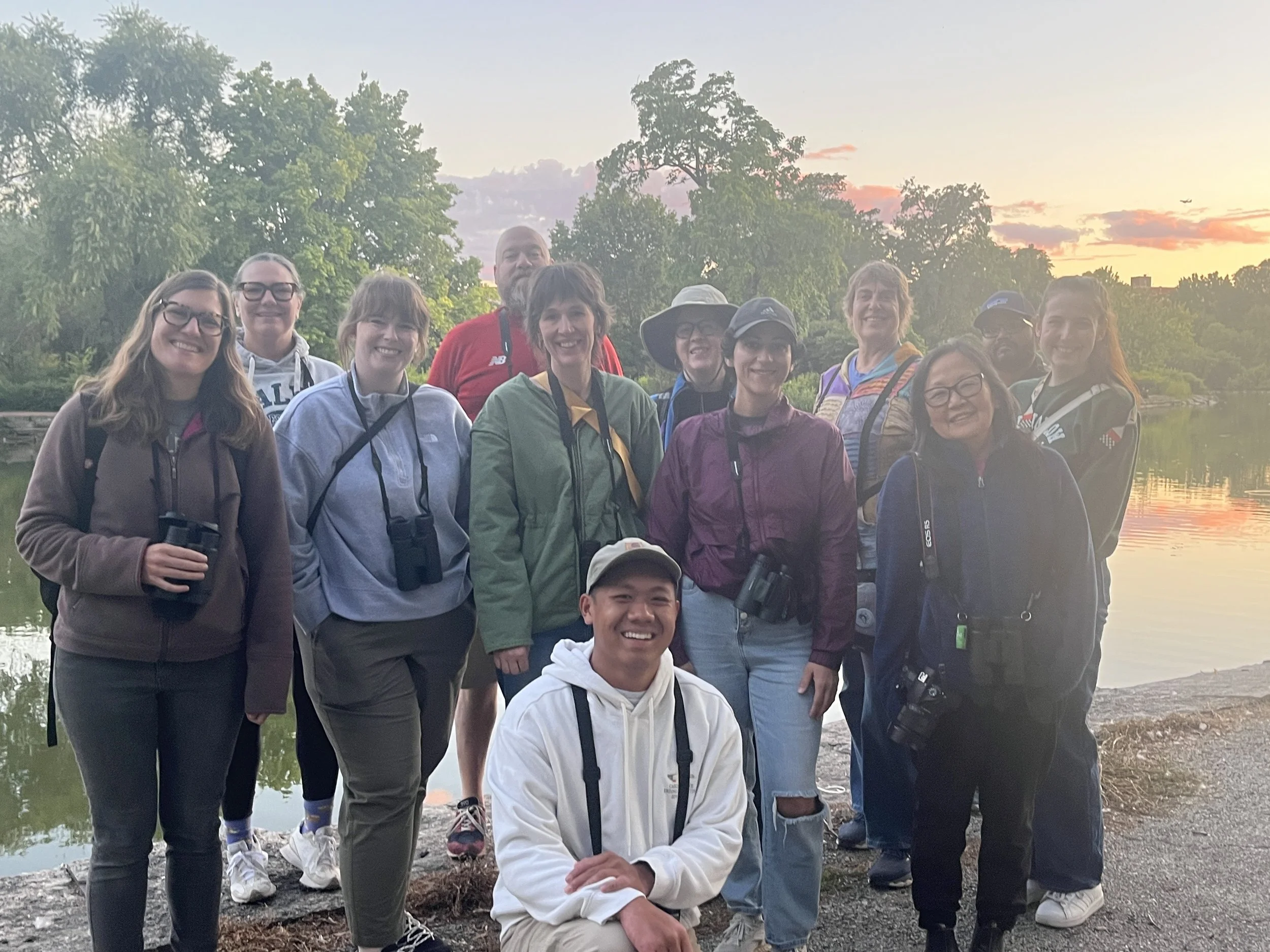 Twelve people with binoculars smiling at camera, with lagoon and a colorful sunset sky behind.