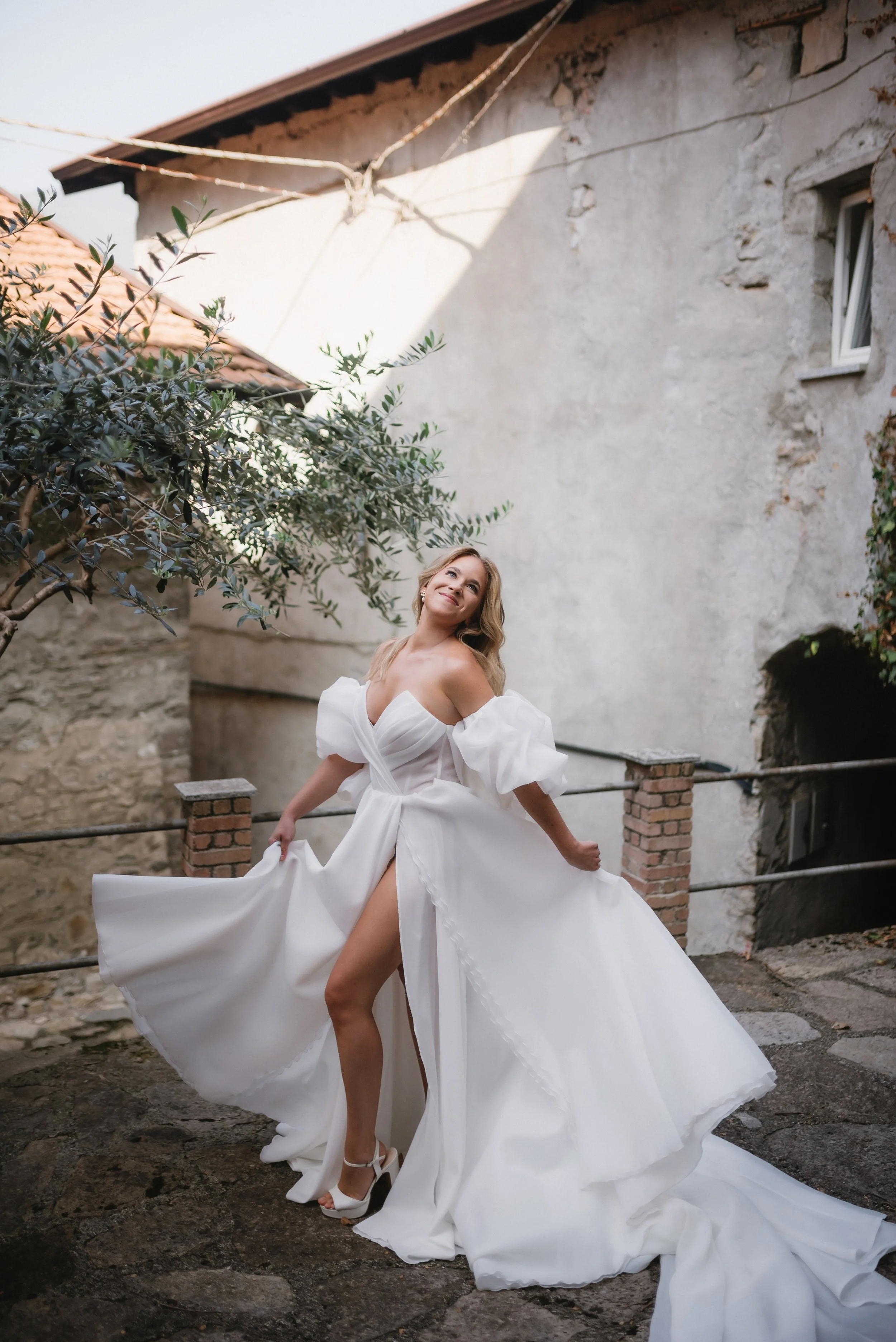 A woman in a white wedding dress with puffy sleeves and a high slit, standing outdoors on a stone path, smiling and holding her dress.