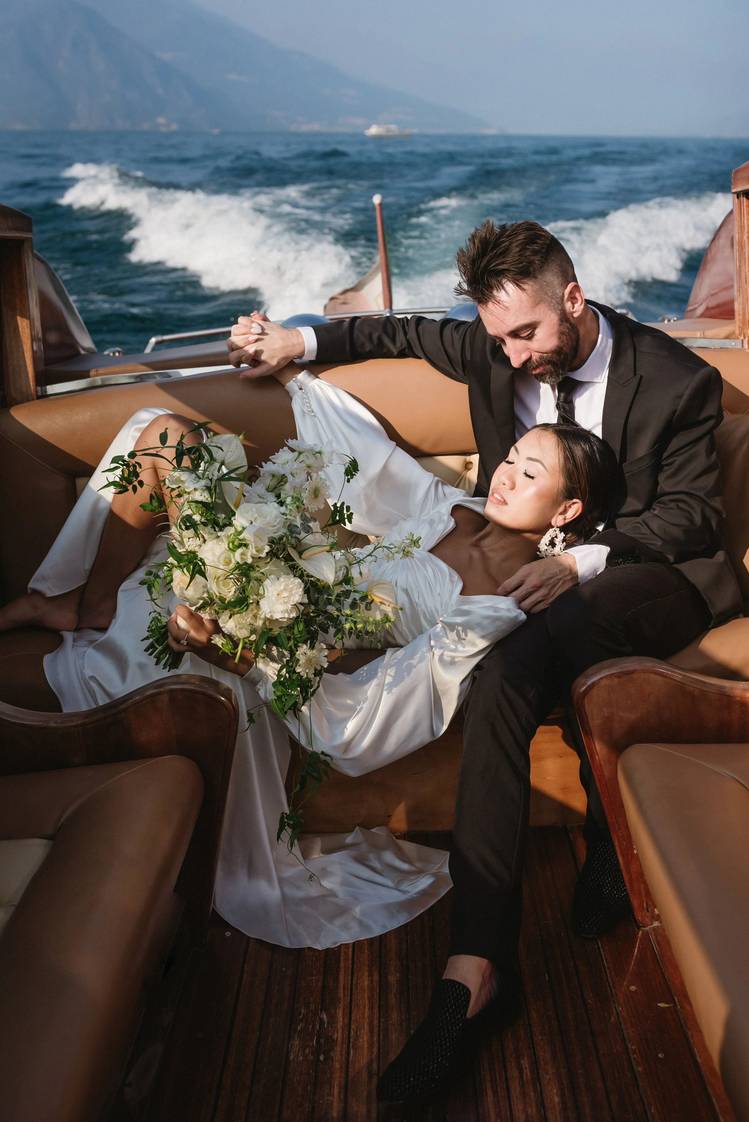 A bride and groom in wedding attire relaxing on a boat with water and mountains in the background.