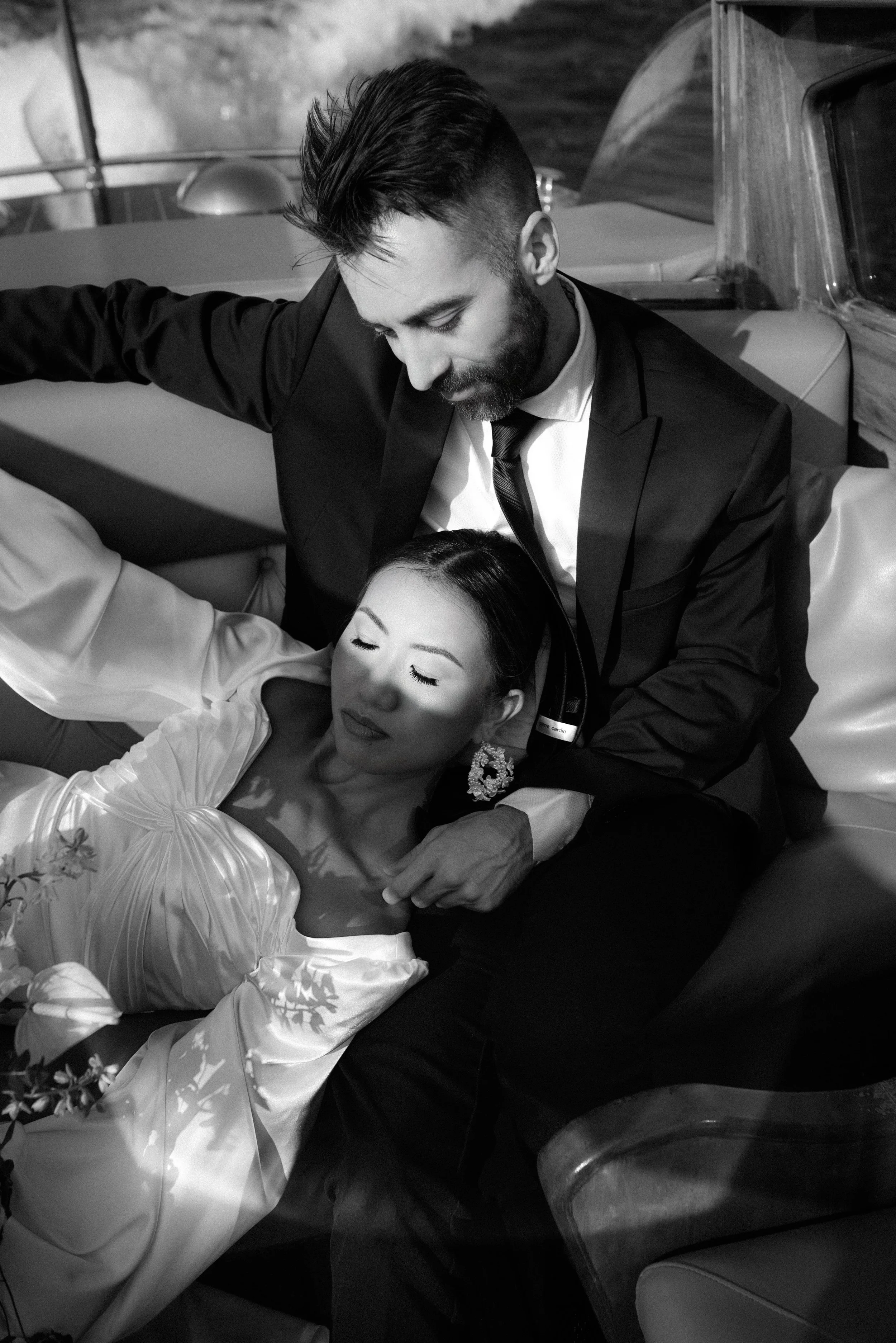 A black and white photo of a man in a suit and woman in a wedding dress resting together on a vintage vehicle, with the woman sleeping and the man looking down at her.