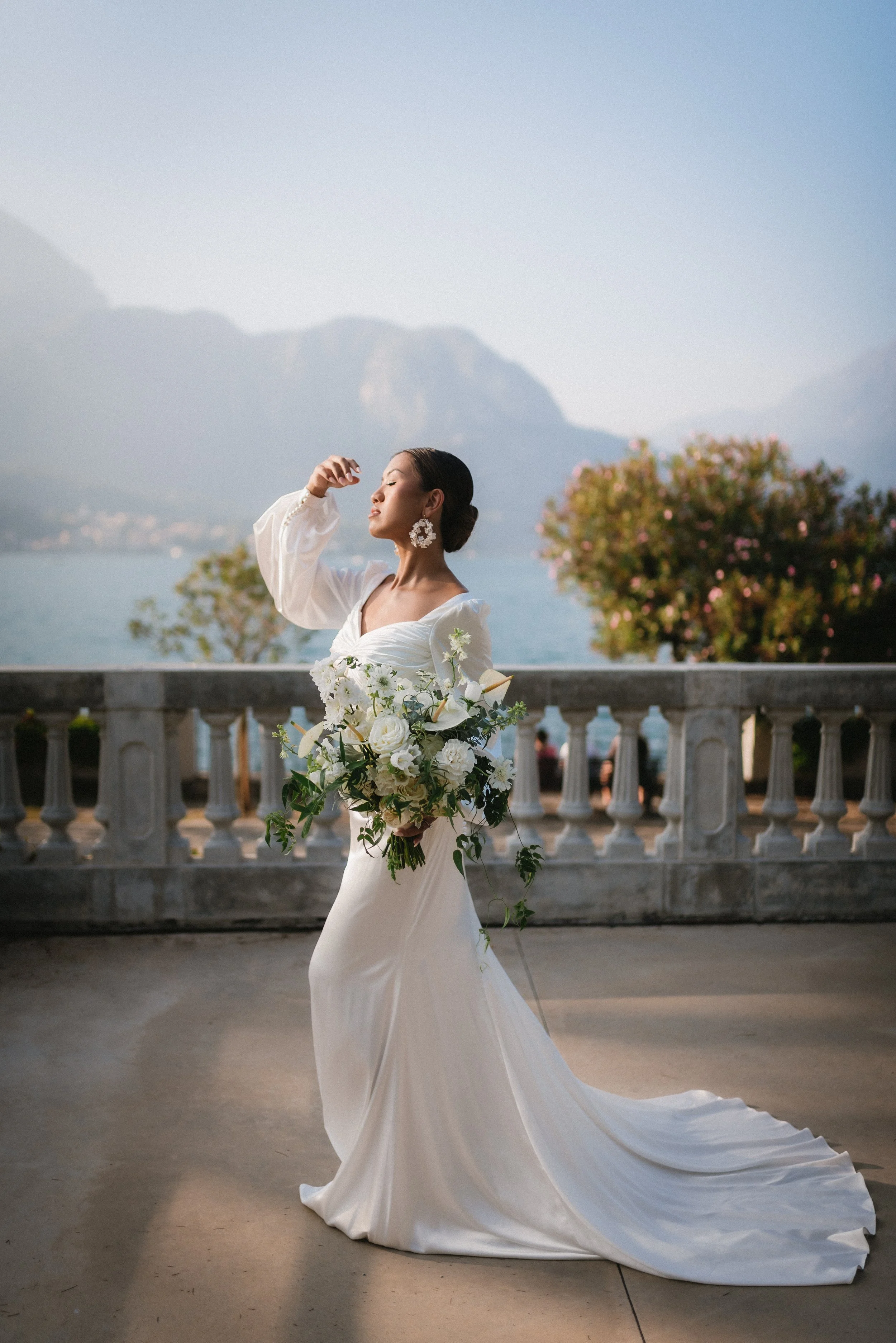 Bride in a white wedding dress holding a bouquet, standing outdoors with a scenic backdrop of mountains and a lake.