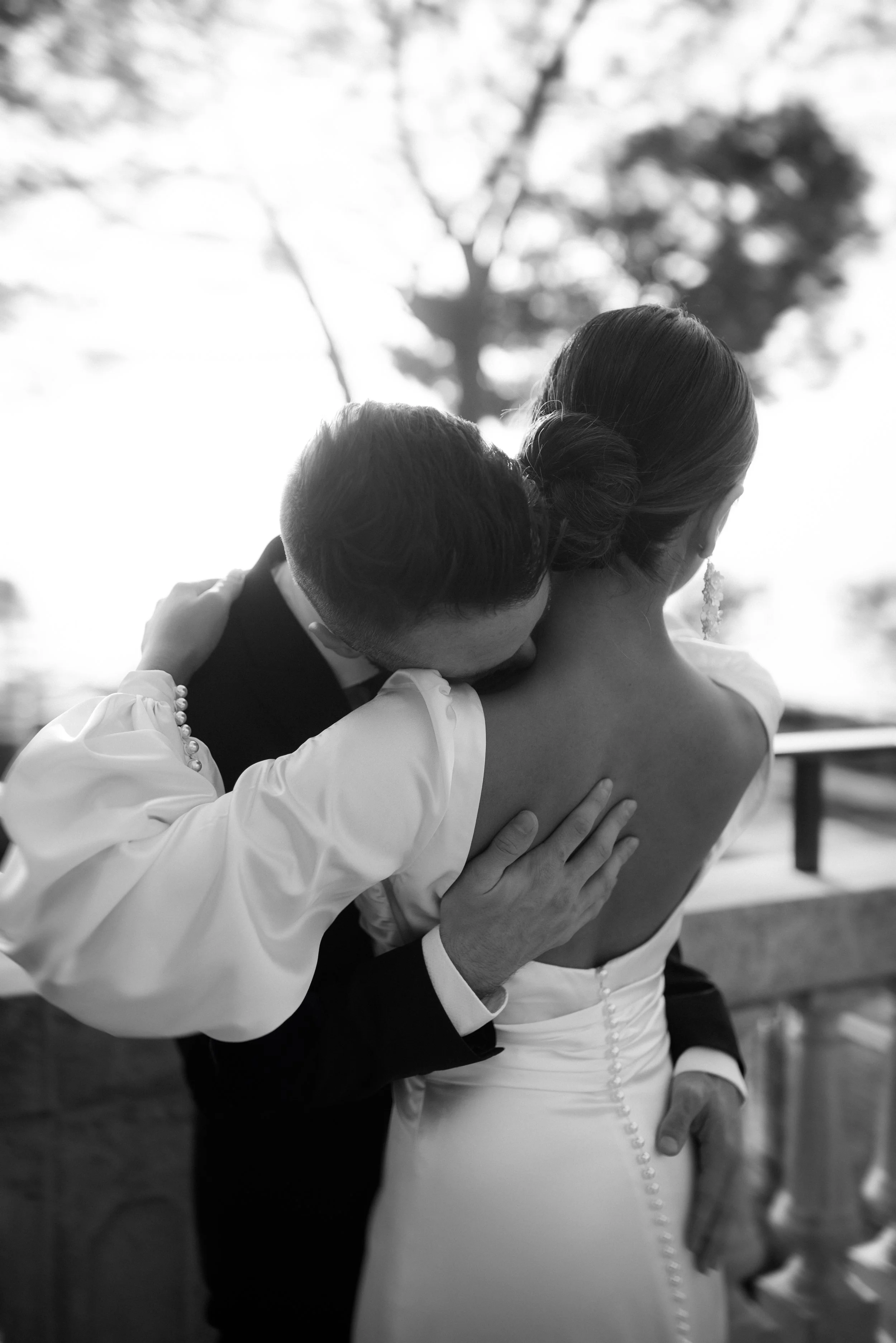 A black-and-white photo of a couple on their wedding day, with the man kissing the woman's neck as she faces away, dressed in a strapless wedding gown.