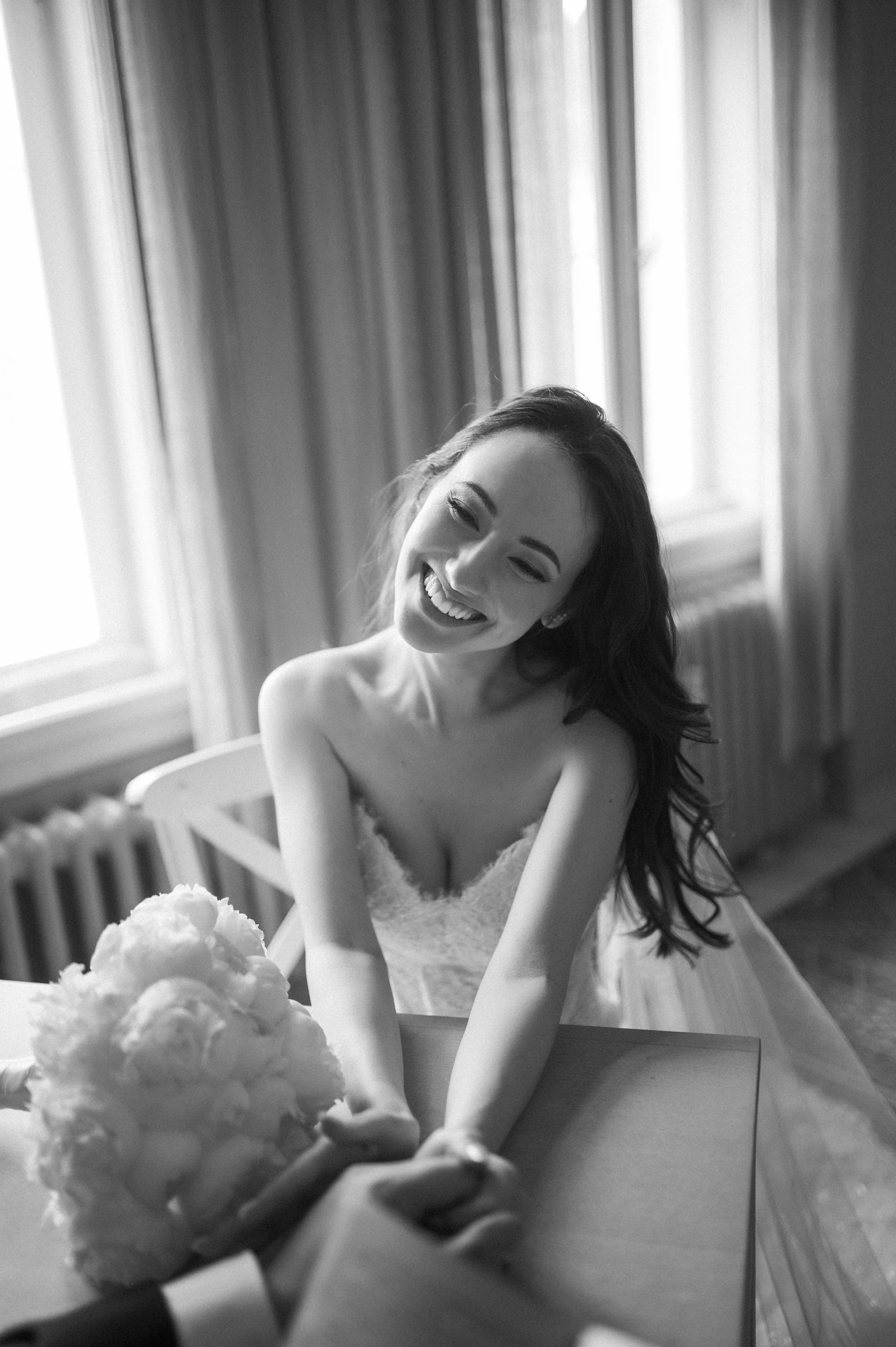 Black and white photo of a smiling bride in a strapless wedding dress holding hands with someone across a table. A bouquet of flowers is on the table.