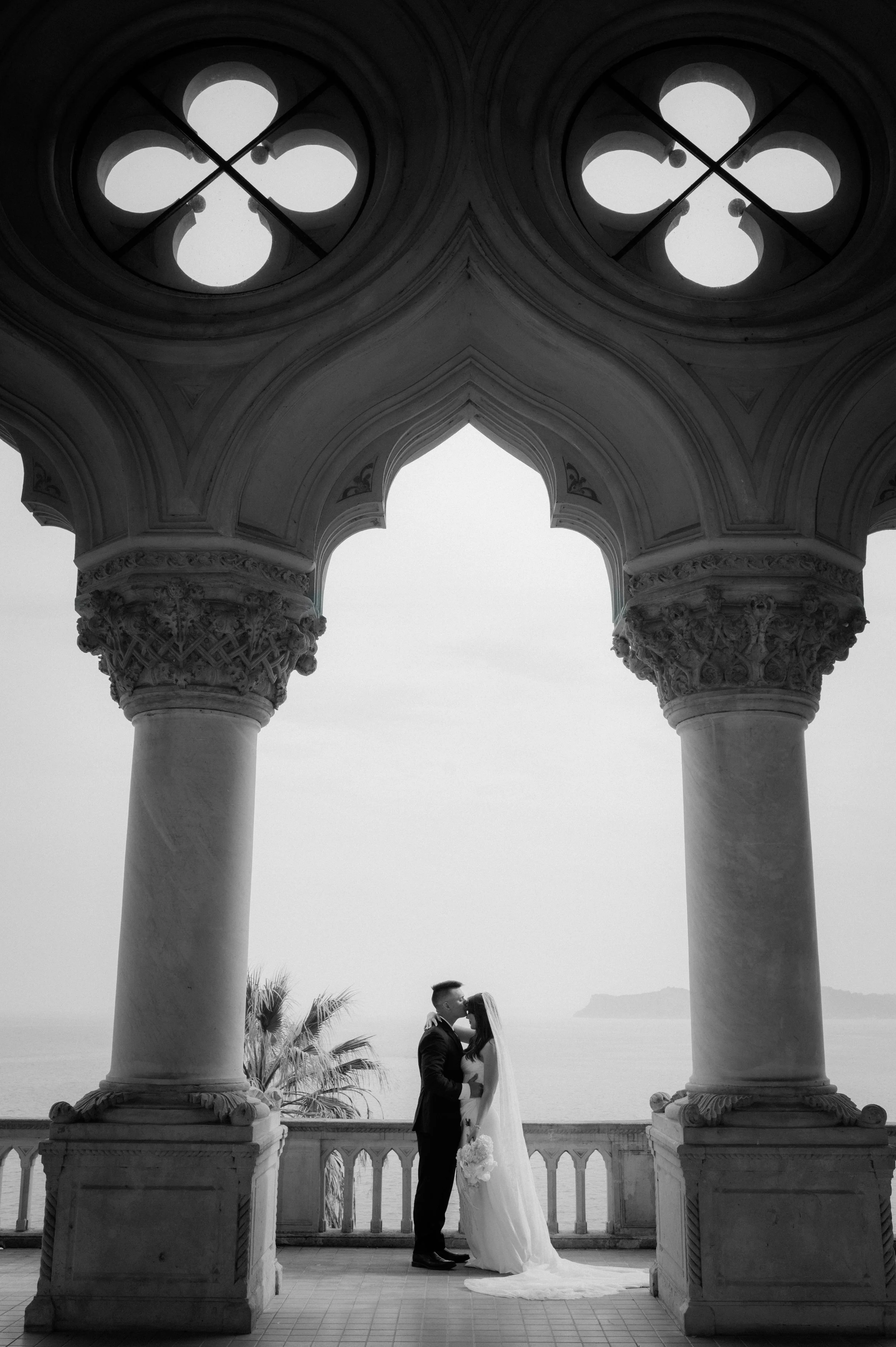 A black-and-white photo of a bride and groom kissing under an ornate archway with columns, overlooking the ocean.