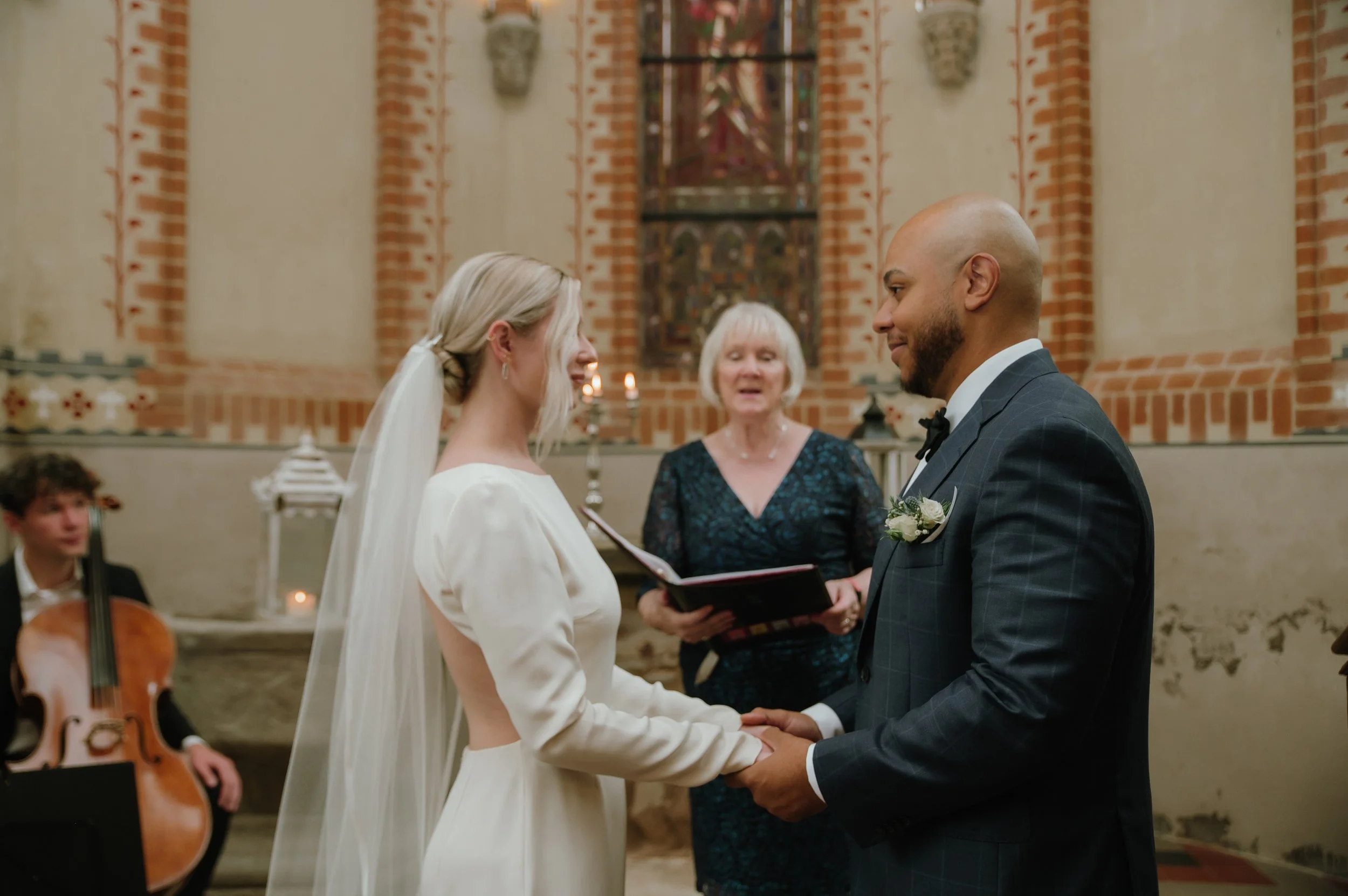 Bride and groom holding hands during a wedding ceremony, with an officiant and cellist in the background.