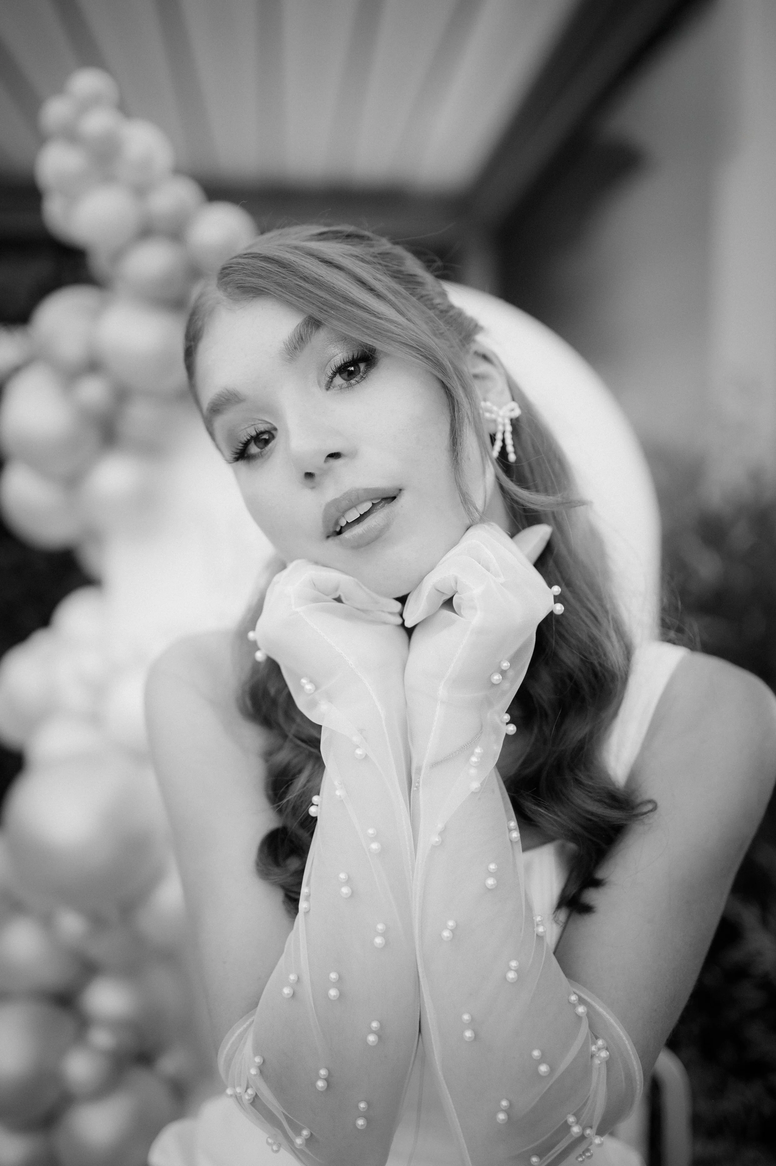 Black and white photo of a woman with wavy hair, wearing earrings and sheer gloves with pearl accents, posing with her chin resting on her hands, in front of a blurred background with balloons.