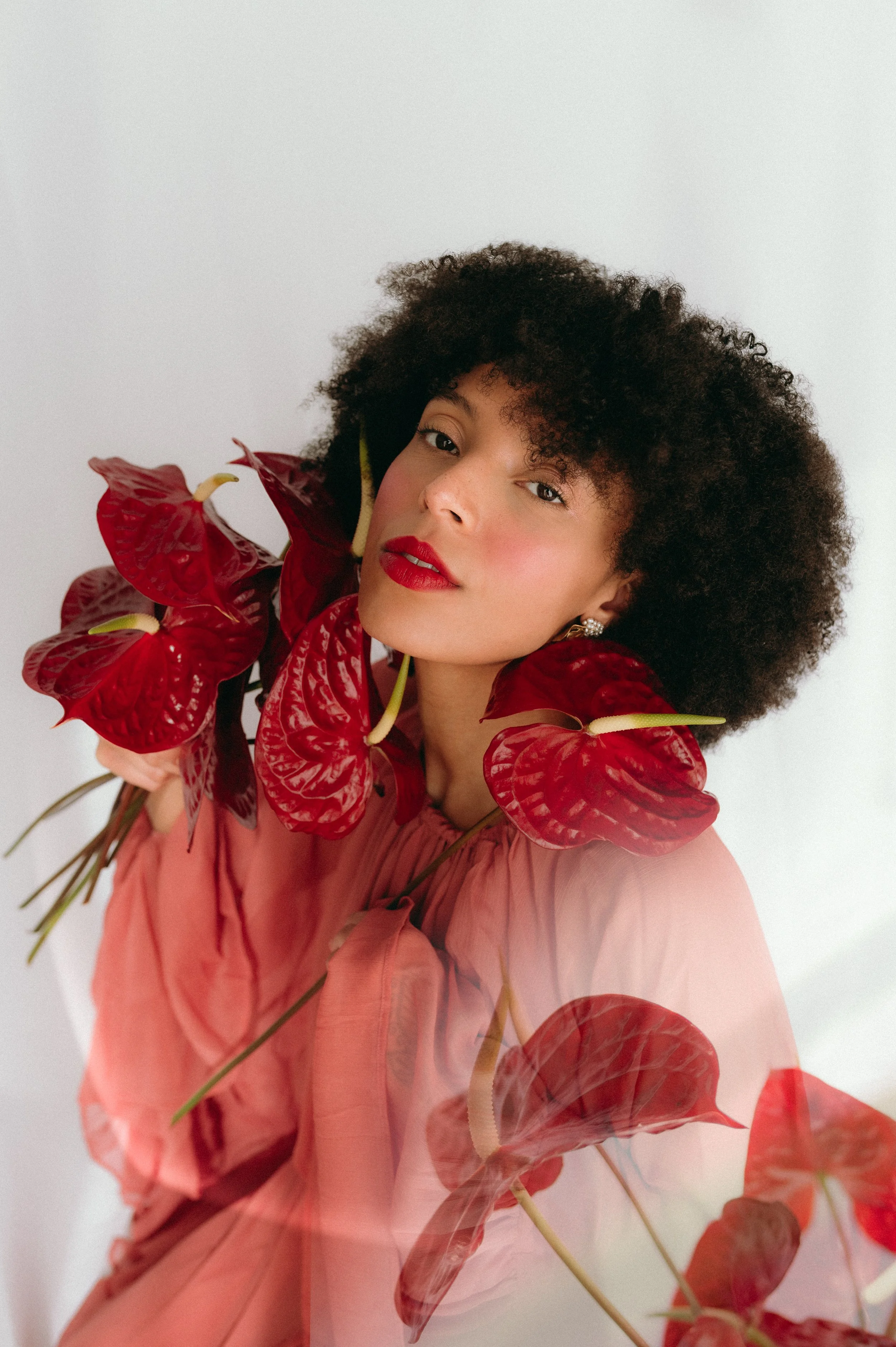 A woman with curly dark hair and red lipstick looks at the camera, surrounded by large red anthurium flowers, against a plain white background.
