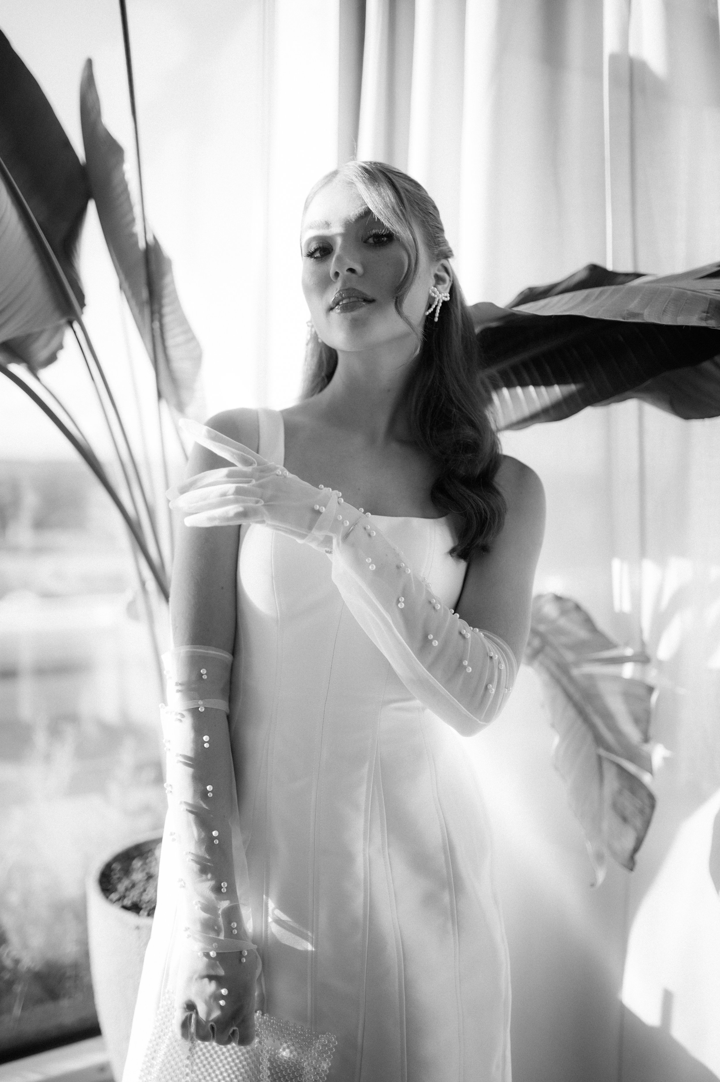 A woman in a white dress with long sheer gloves, standing indoors near large decorative leaves, in a black and white photo.