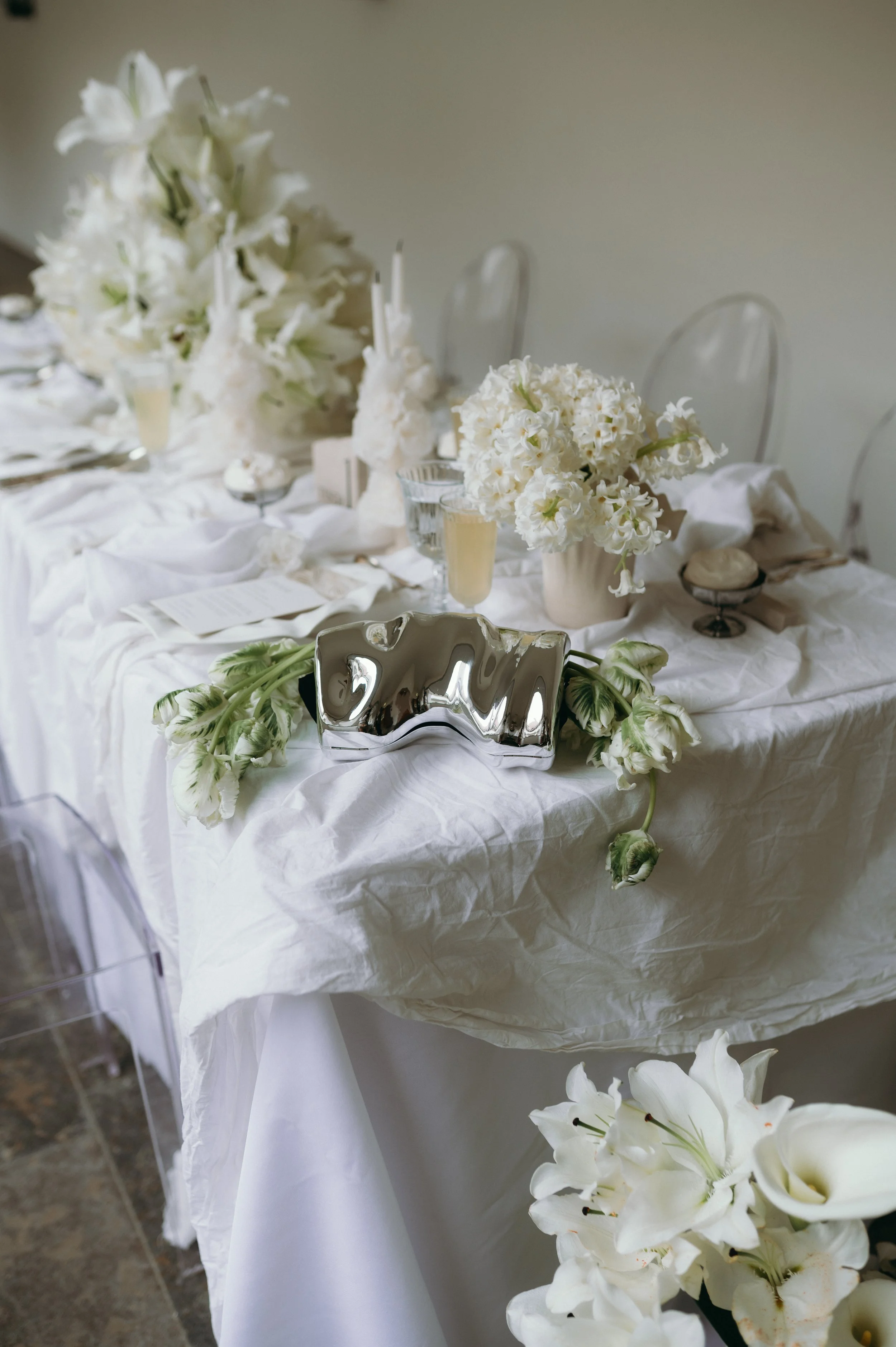 Elegant table setting with white flowers, candles, and a shiny metallic clutch, decorated for a sophisticated event or celebration.