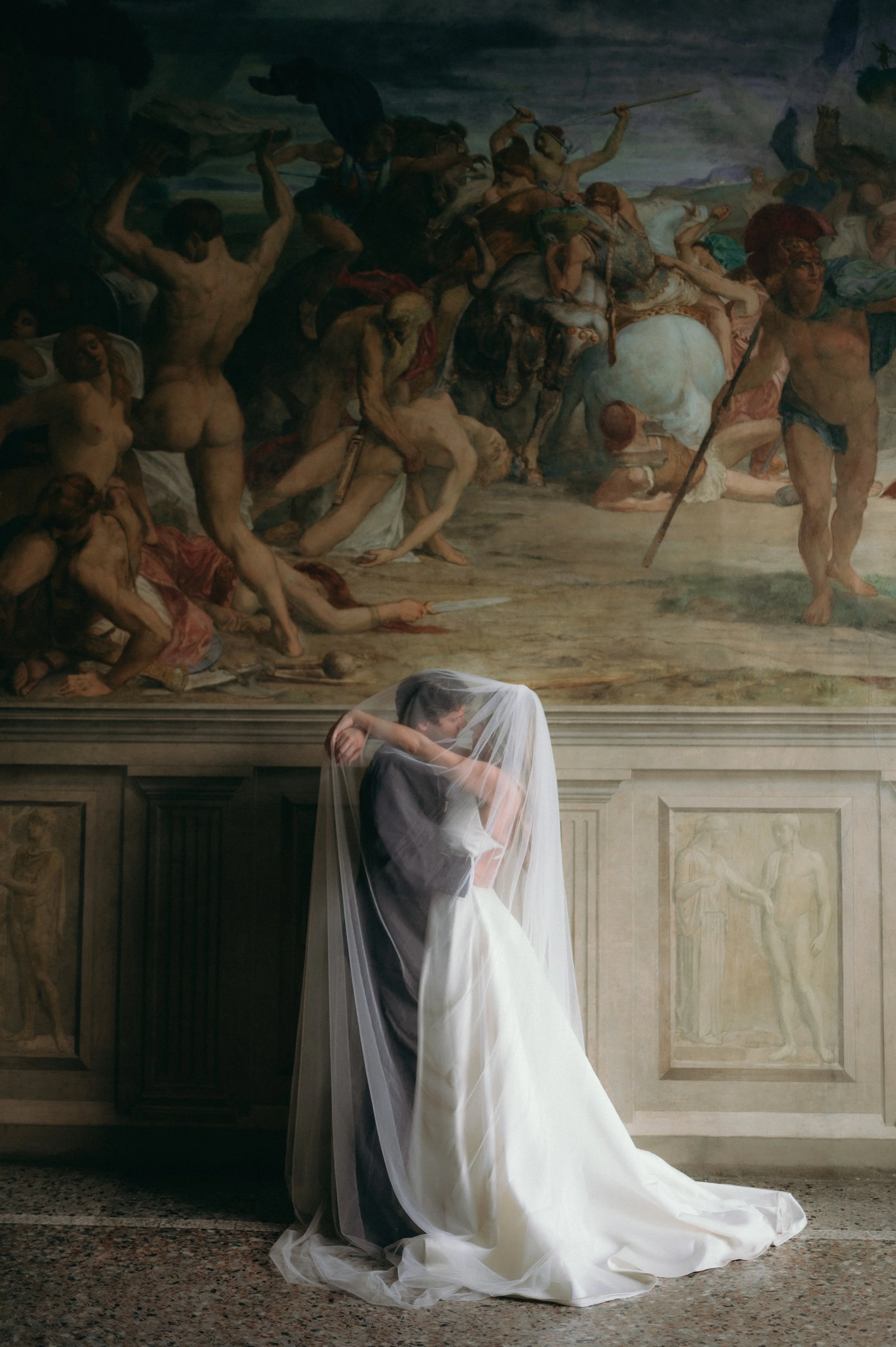 A bride and groom kissing behind a veil in front of a large classical painting depicting a historical or mythological scene.