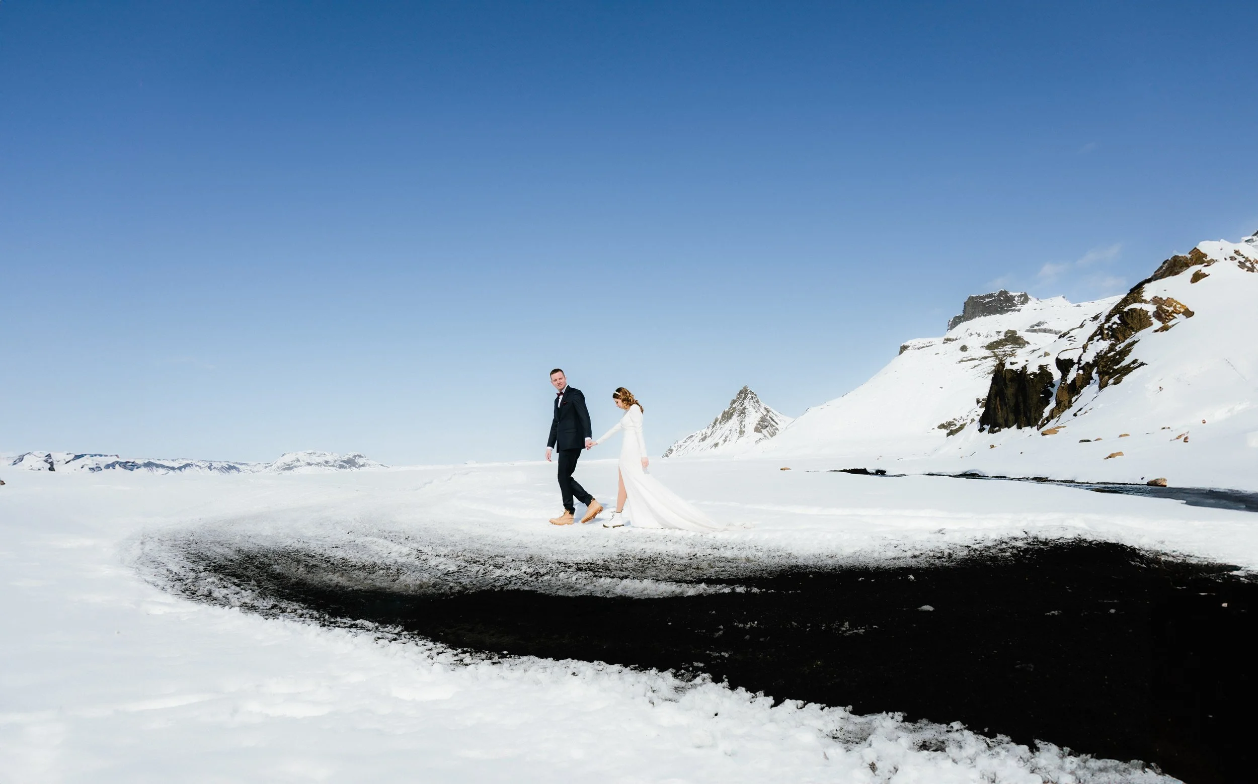A bride and groom holding hands and walking on a snowy landscape with mountains in the background on a clear day.