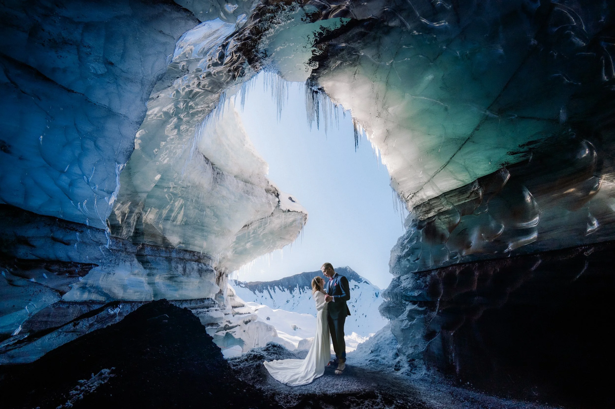 Couple standing in a glacier cave with ice formations and icicles, wearing wedding attire, and surrounded by snow and mountains.