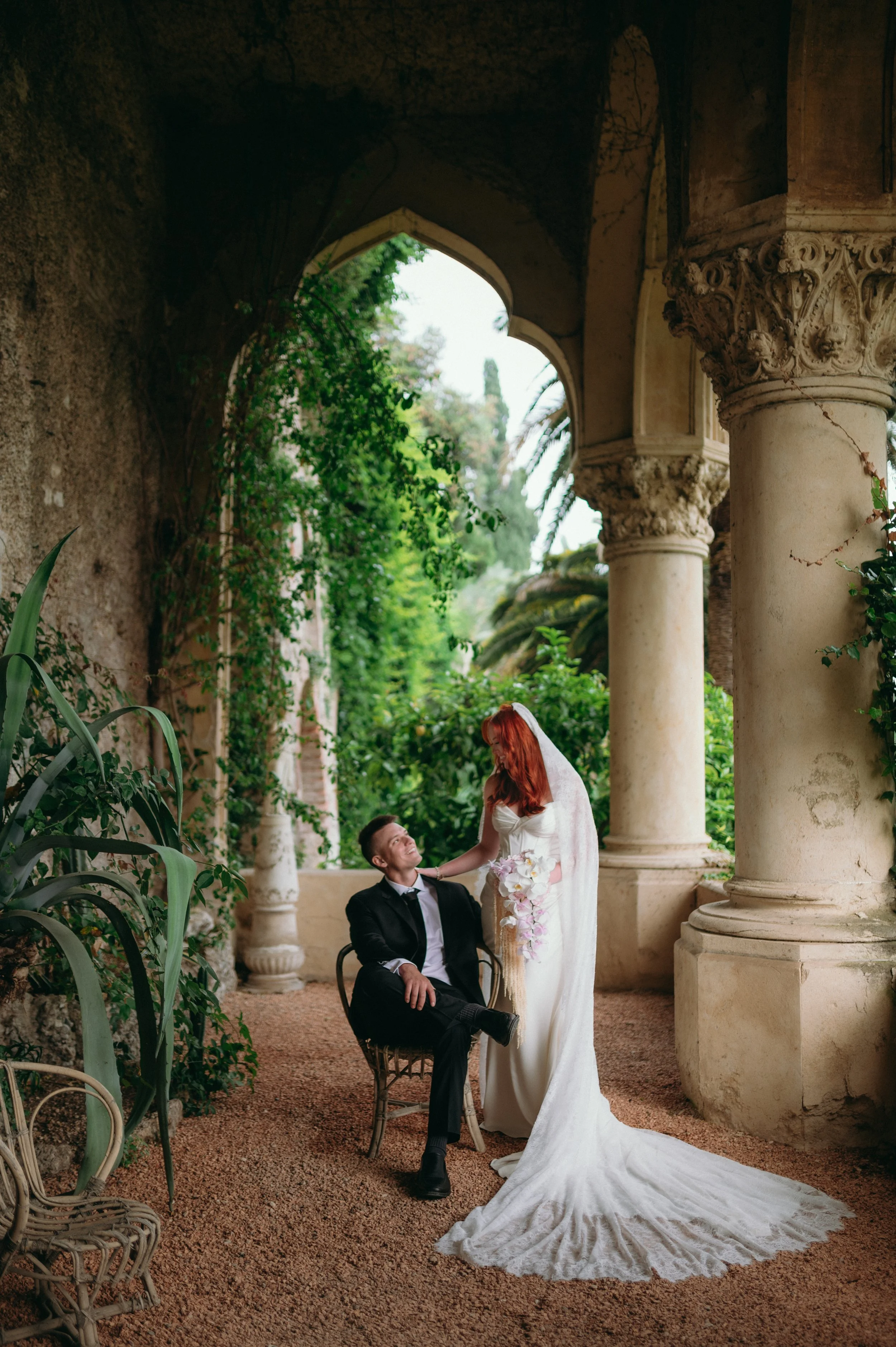 A bride and groom pose in an outdoor, historic architectural setting with columns and greenery. The bride is dressed in a white wedding gown with a long train and veil, holding a bouquet of flowers, and is looking at the groom. The groom is seated in