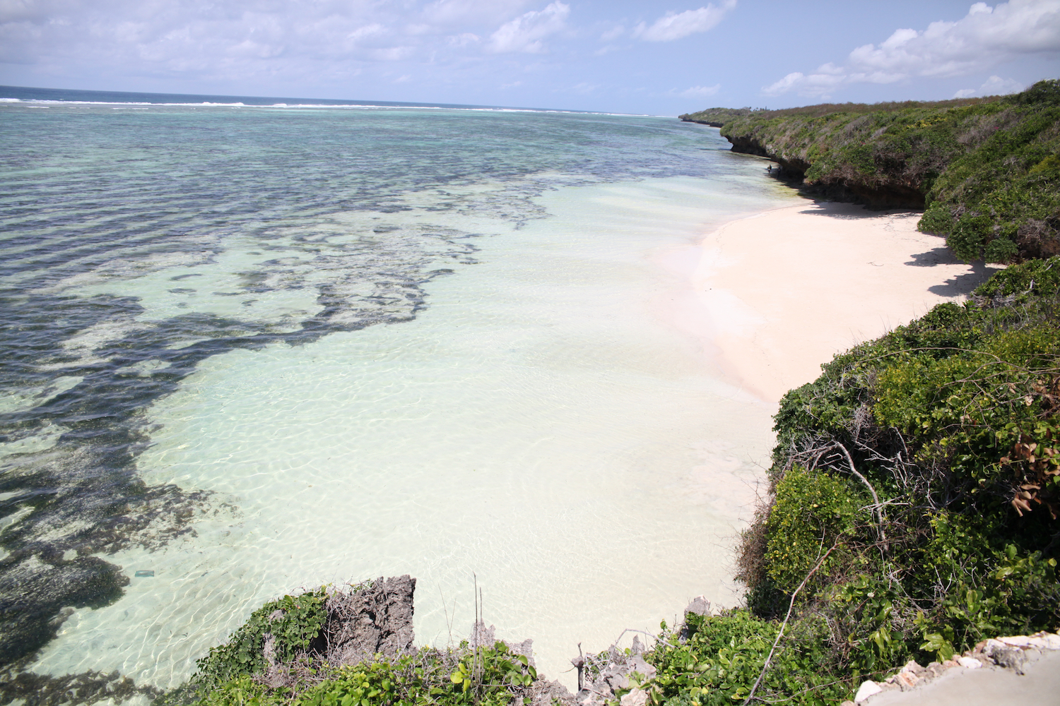 A serene beach scene with clear turquoise water, white sand, and rocky cliffs. The lush green vegetation adds contrast, under a bright, partly cloudy sky.
